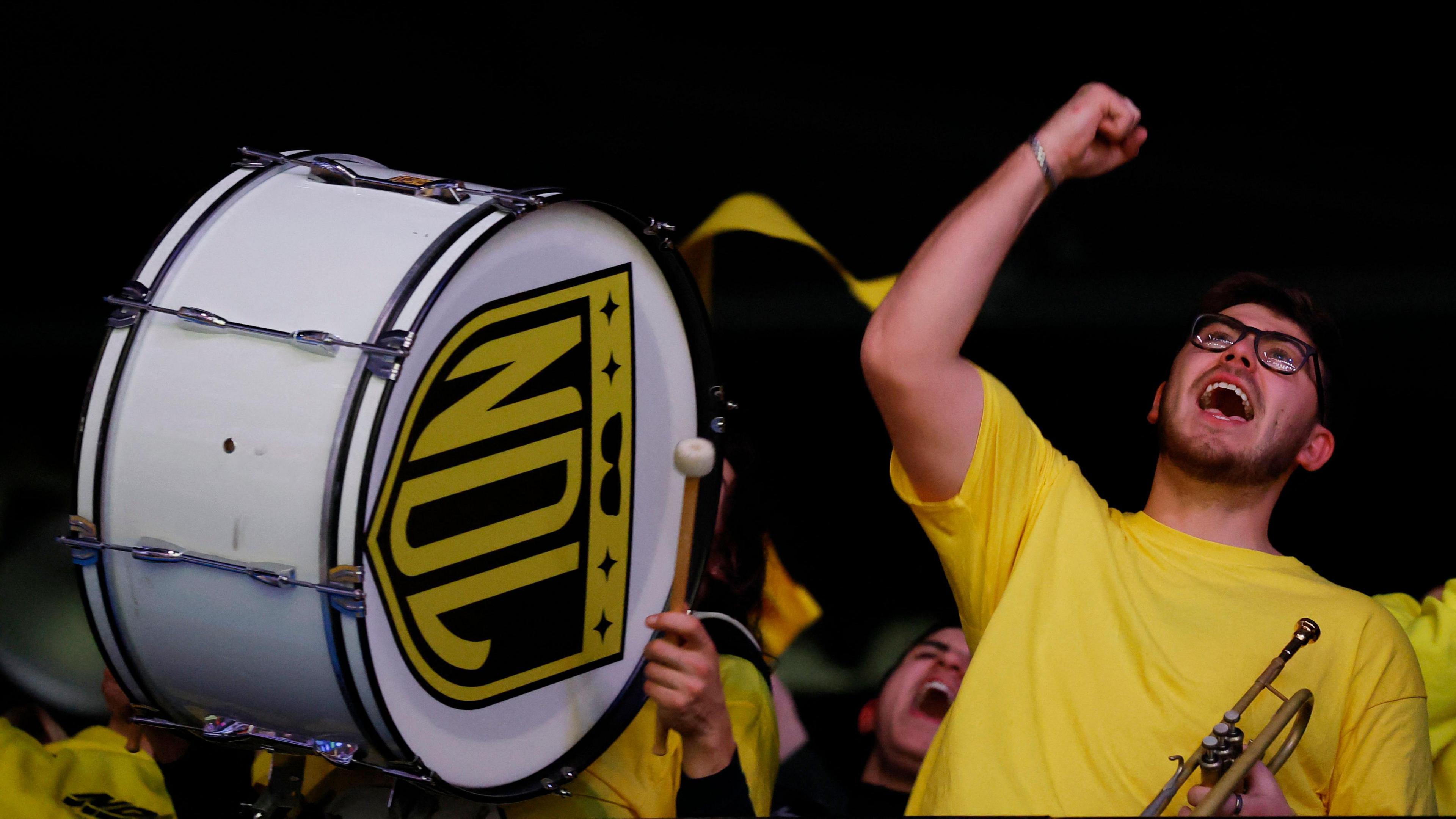 A person wearing a yellow t-shirt holds a trumpet and is standing next to a drum
