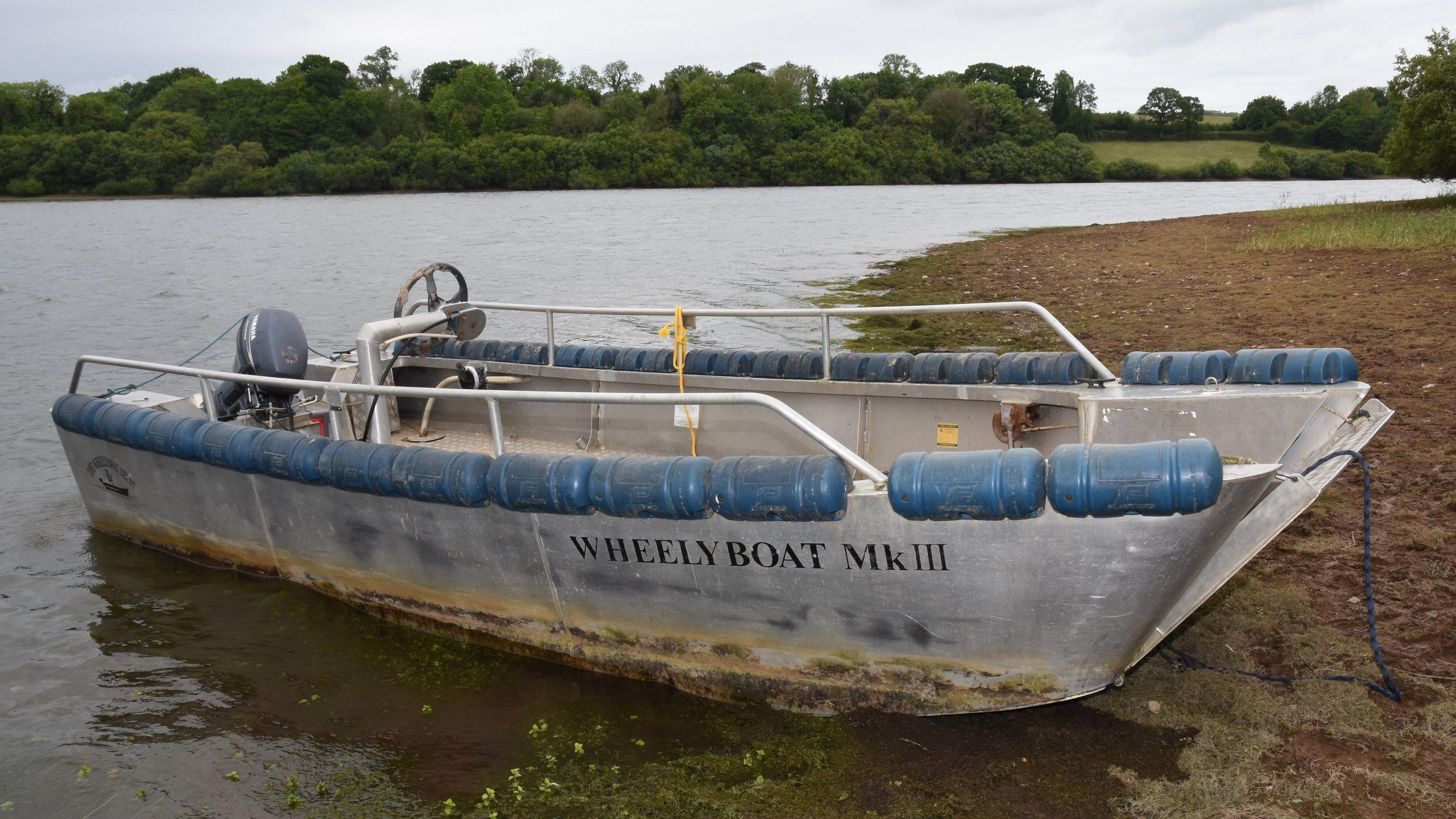 The Wheelyboat Mk III is tied to a muddy foreshore with the lake behind it. The boat has water marks across its hull.