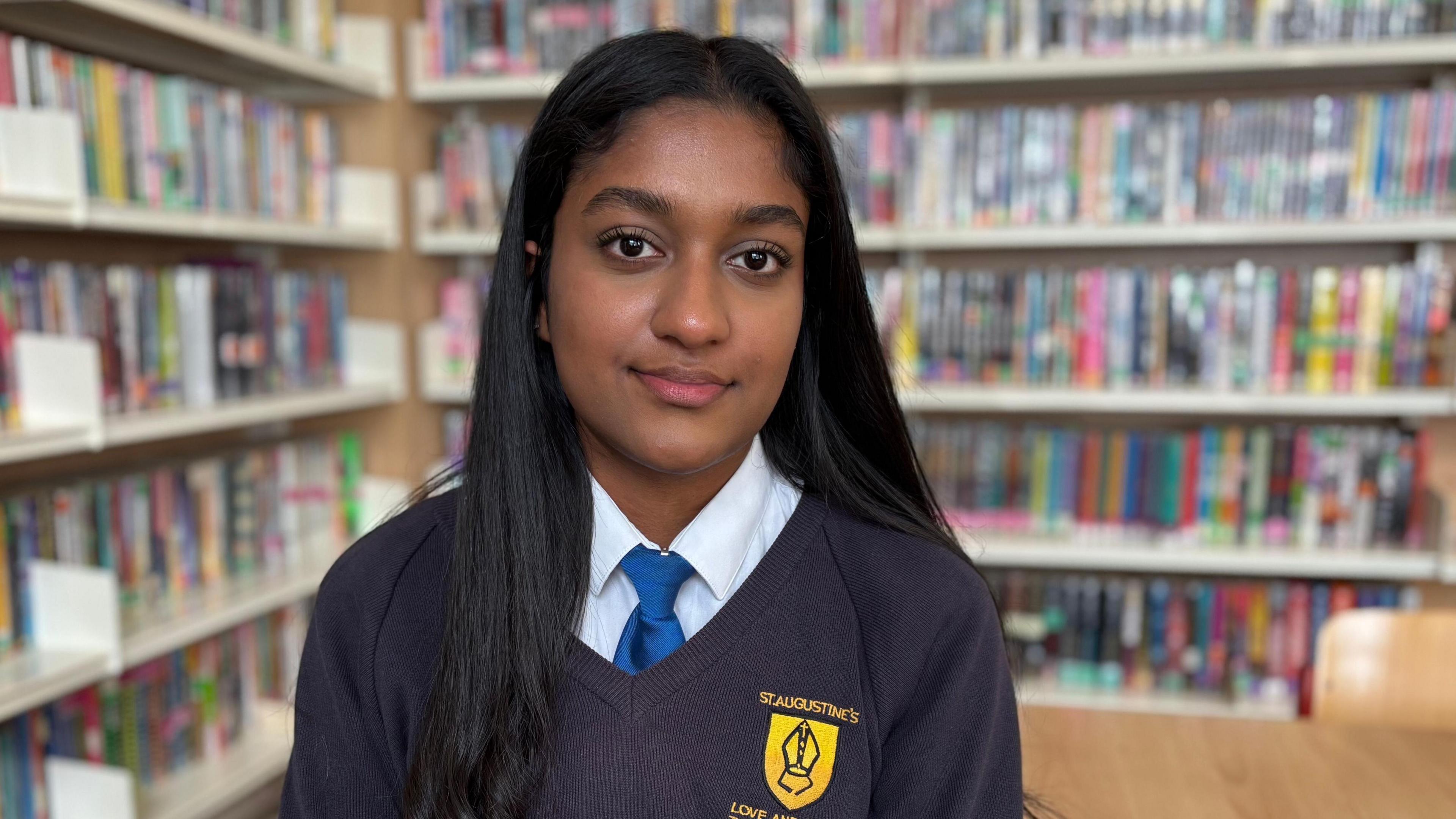 Jiya has long dark hair which is curled at the end. She wears a branded navy blue v-neck school jumper, a white shirt, and a bright blue tie. She is sat down next to a table and is smiling directly in to the camera. Behind her, out of focus, are shelves and shelves of books. 