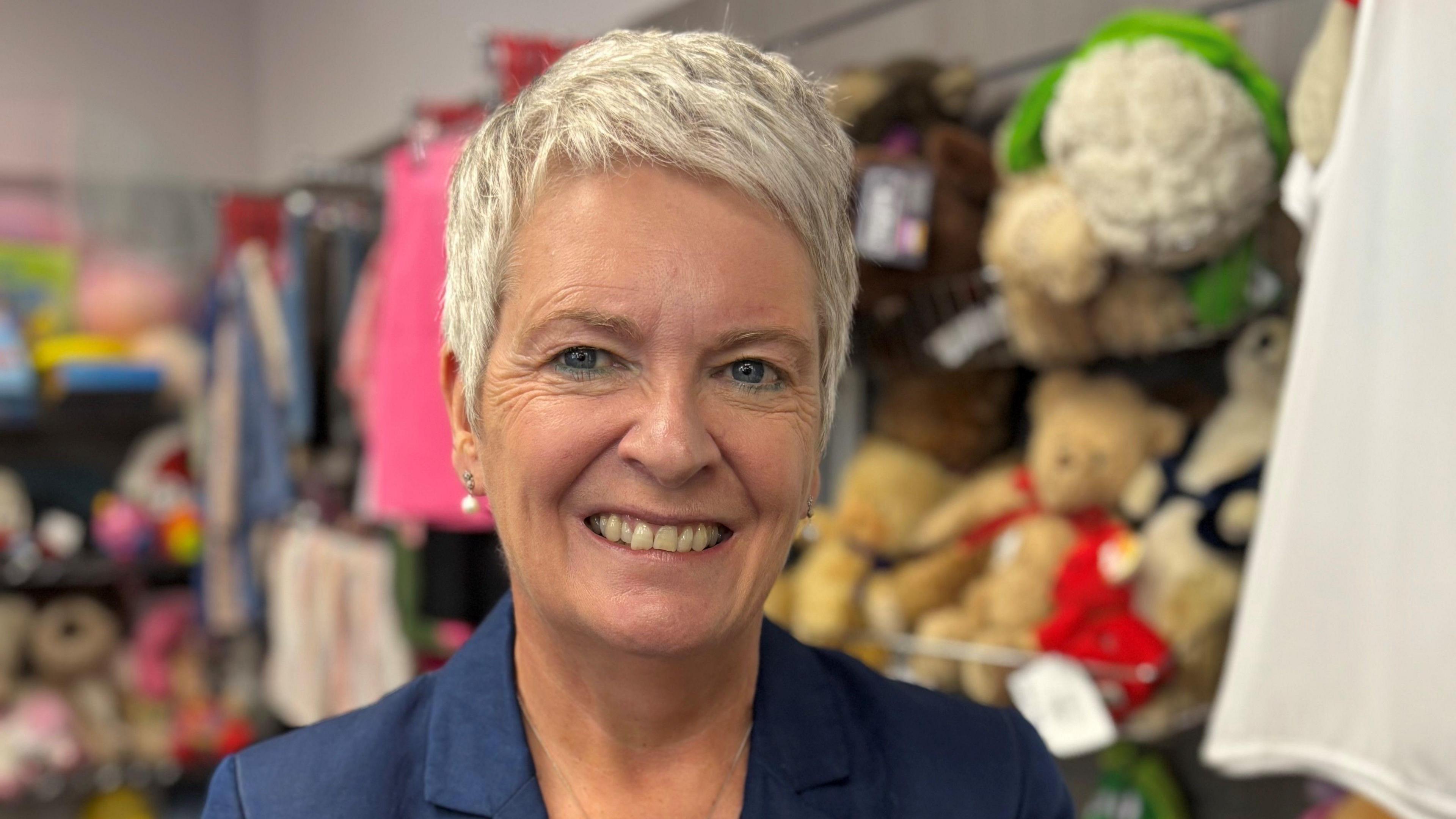Report author Edel Harris - a smiling woman with short blonde hair, wearing a blue blazer and drop pearl earrings - stands in front of shelves of soft toys.