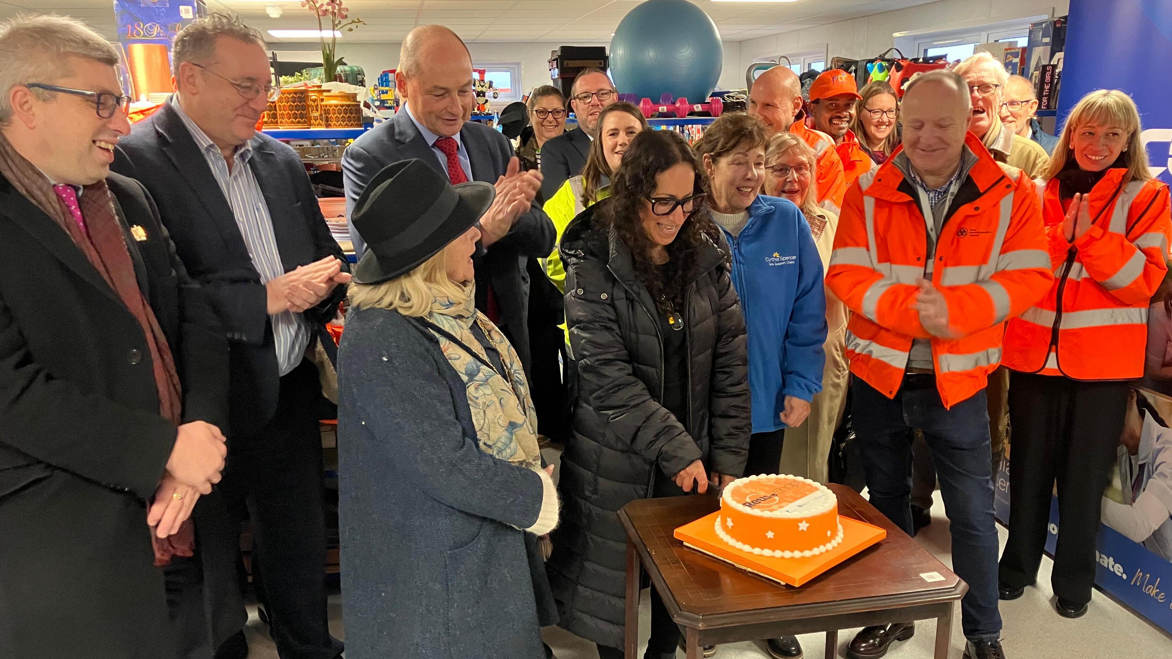 Lots of people standing around an orange cake clapping and smiling. In the background lots of items are on shelves for sale.
