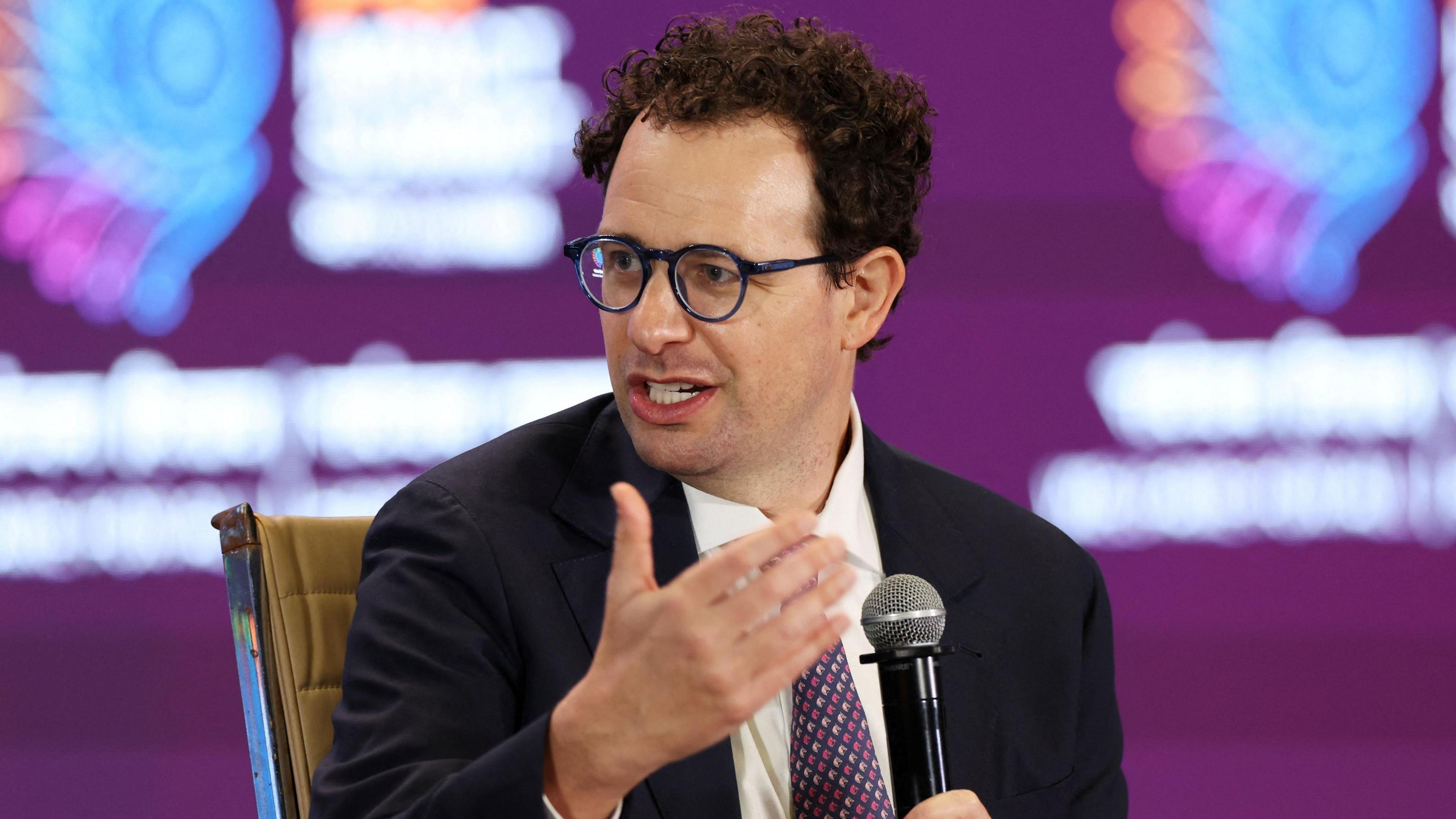 CEO of Anthropic Dario Amodei, addresses the gathering at the AI Impact Summit, in New Delhi, India. He wears glasses, a dark suit and a purple tie, sitting against a purple background. There is a microphone in his hand as he addresses somebody unseen.