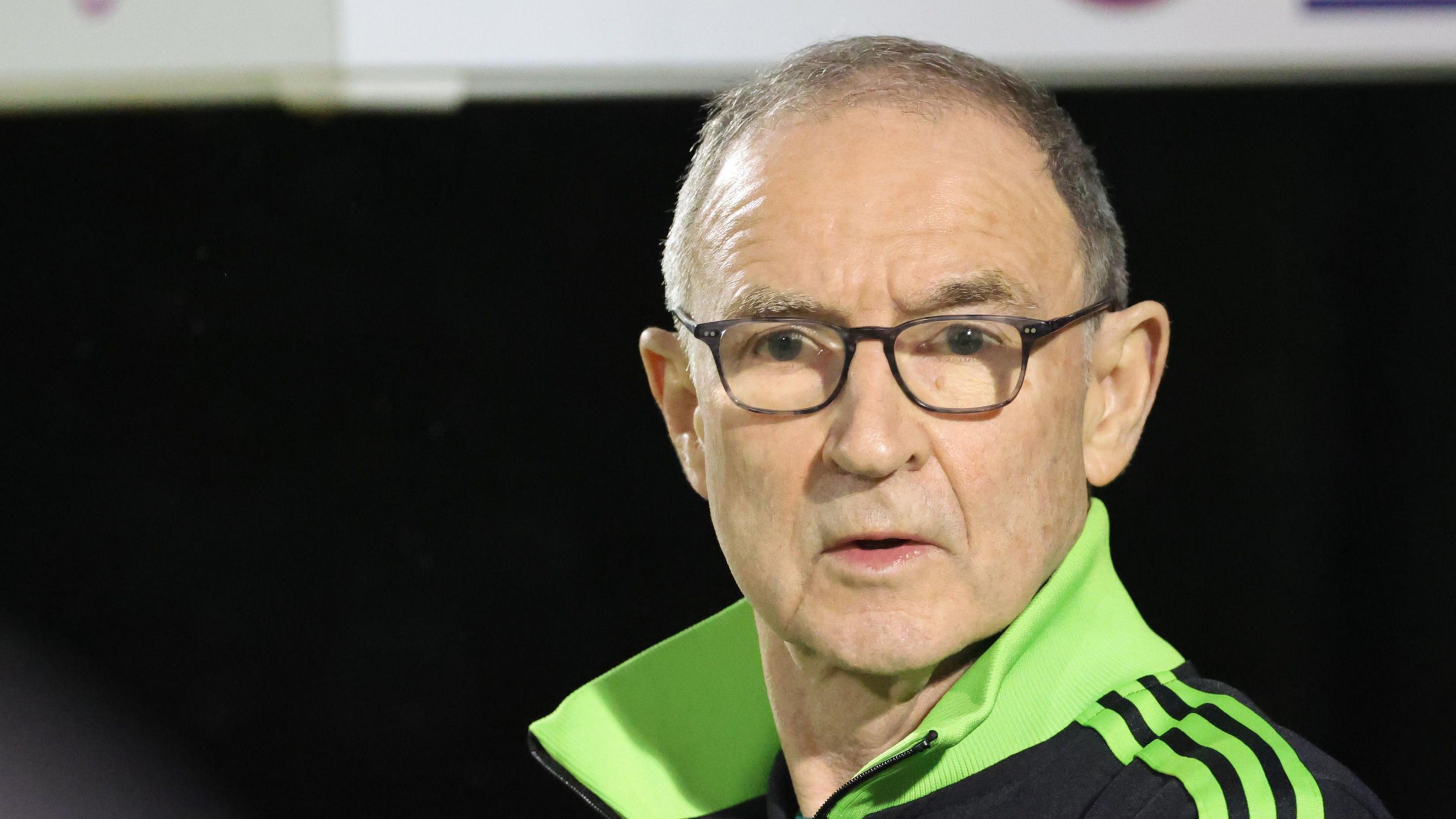 Martin O'Neill is a white man with dark grey hair. He wears glasses and a back and bright green top