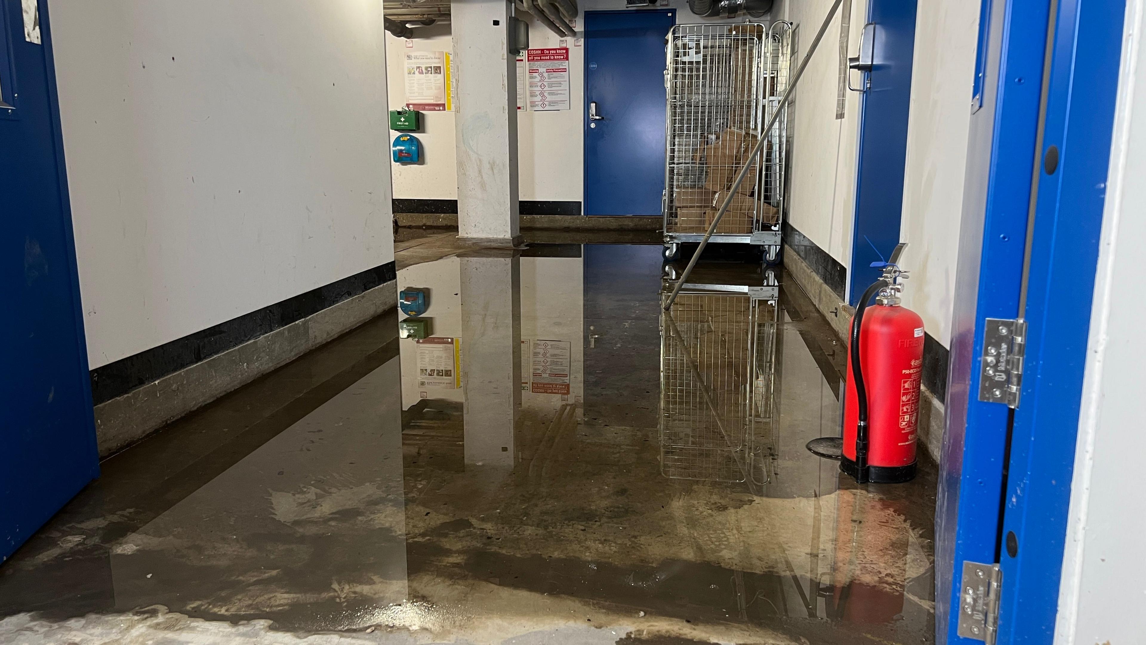 A floor in a hospital covered in a centimetre or two of water. The wall are painted white, with doors painted blue. There is a fire extinguisher on the floor - sitting in the water on one side of the corridor. A trolley cage with what appear to be brown parcels is at the end of the corridor.