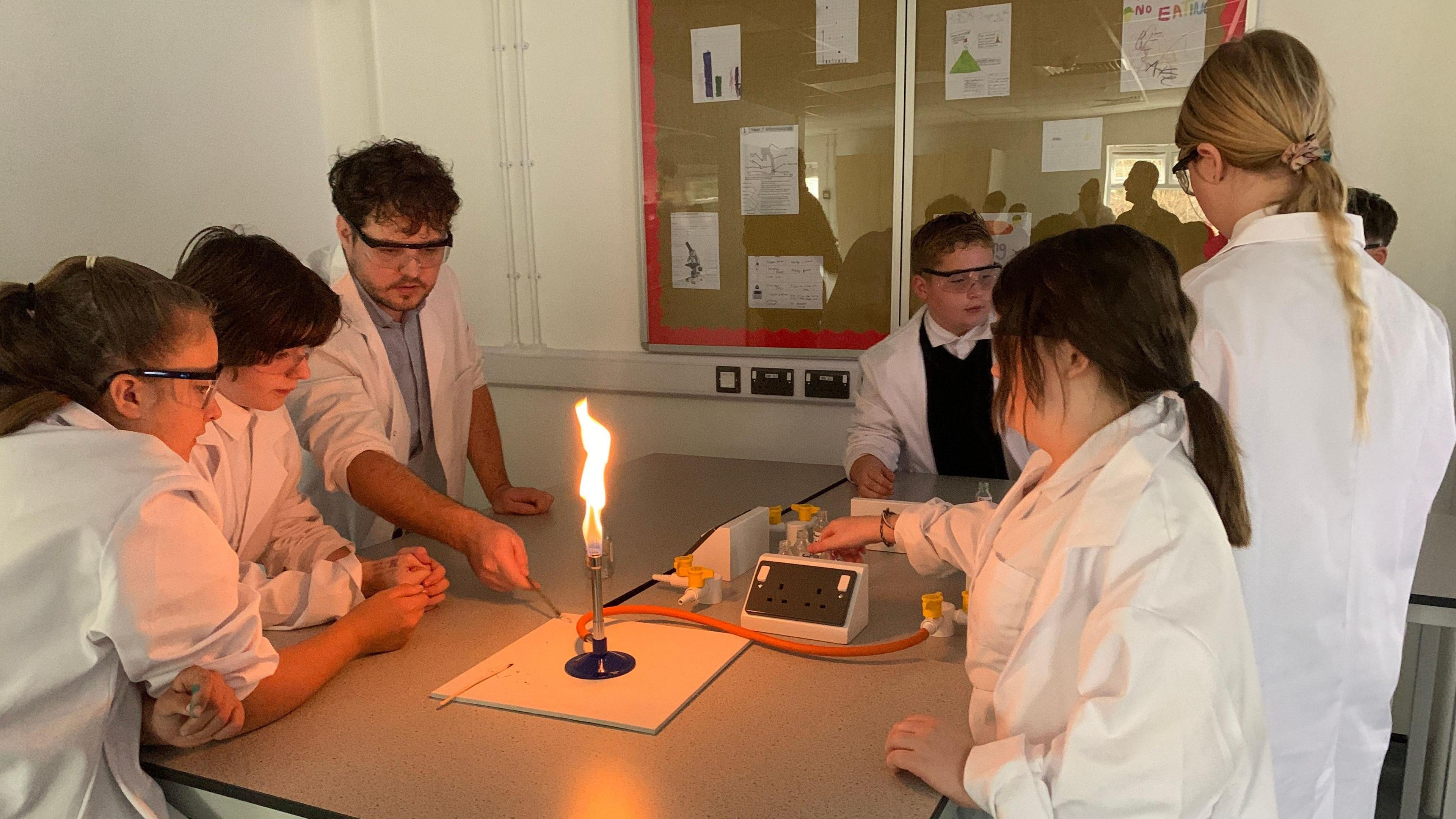 Children gather round a bunsen burner in a classroom for an experiment that a teacher is demonstrating