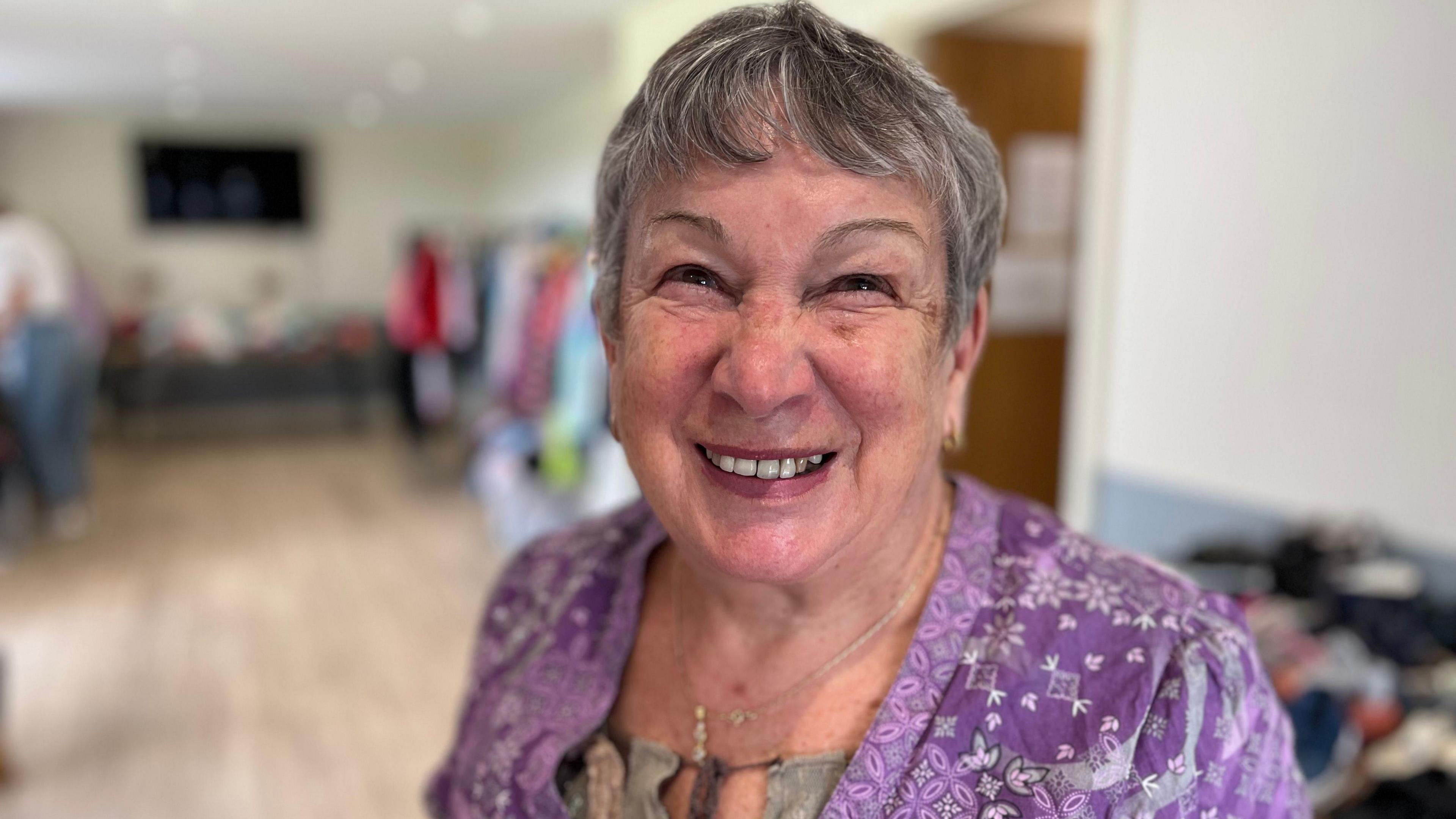 A woman with short grey hair squinting at the camera. She's smiling shopwing her teeth. She's wearing a purple blouse which has a flowery pattern on it.