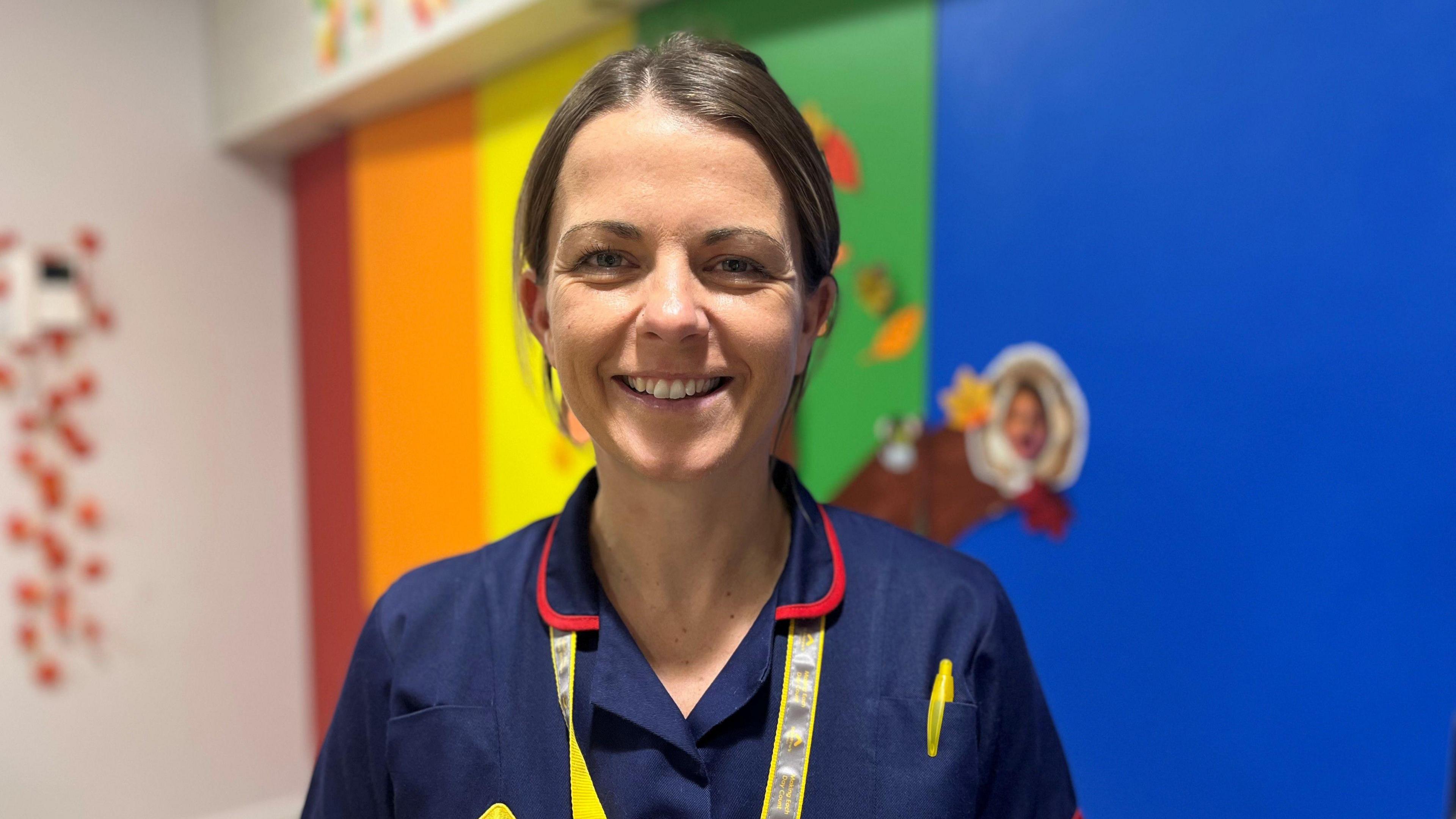 Nurse Lucy Williamson looks directly at the camera and smiles. She is wearing a navy blue nurse's uniform with a red collar, and has a yellow lanyard and yellow pen in a pocket on the right hand side. The background is colourful - a rainbow, painted on one of the walls in the hospice. 