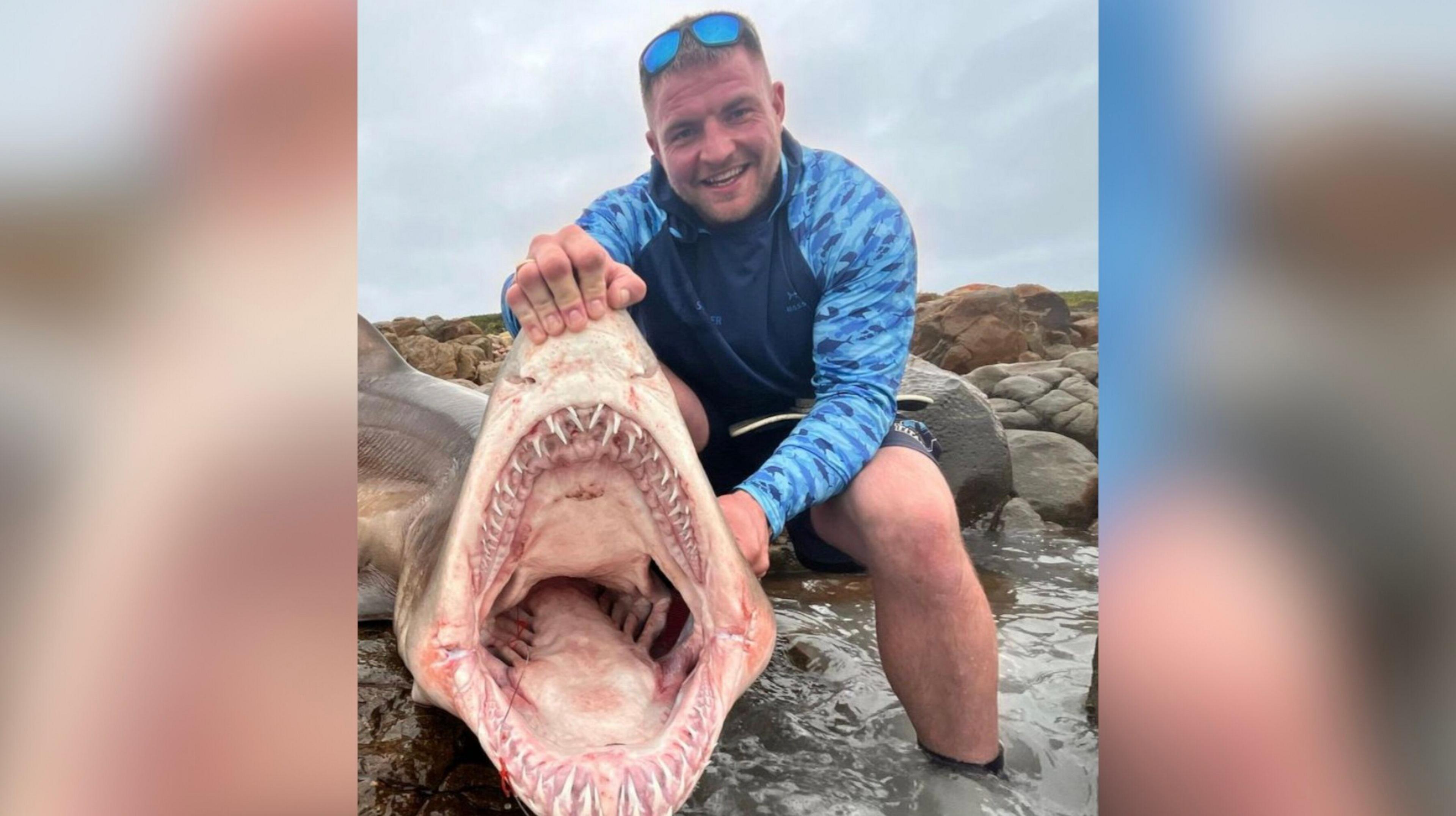 Curtis Miller, holding the jaws open of a ragged tooth shark while standing in shallow water in rocks.