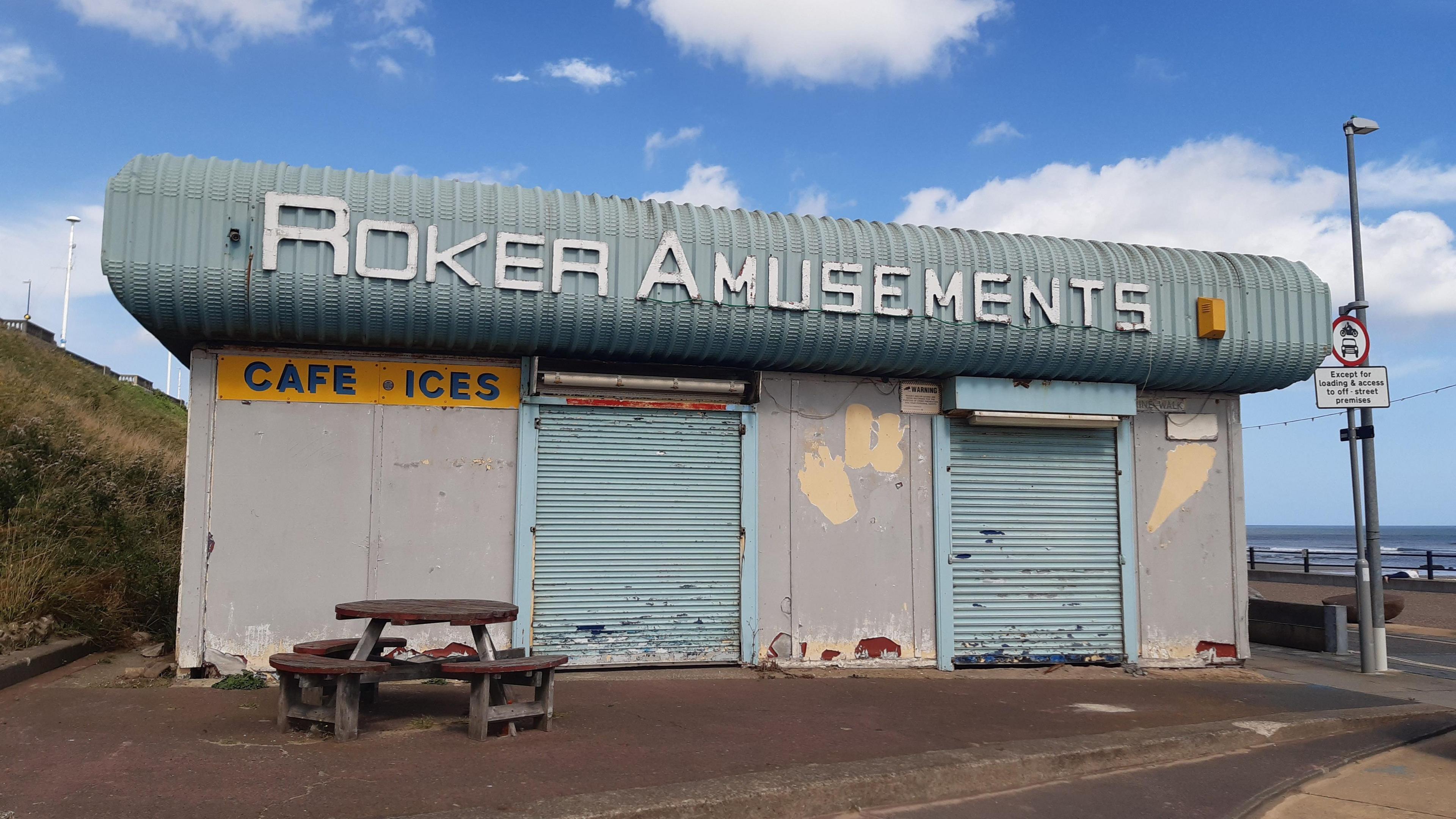 The building is grey and its doors have their shutters down. Paint is peeling off the walls. The roof is rounded and made of corrugated metal. A sign reading Roker Amusements is on the building. A picnic bench sits outside. The sea can be seen to the left in the background.