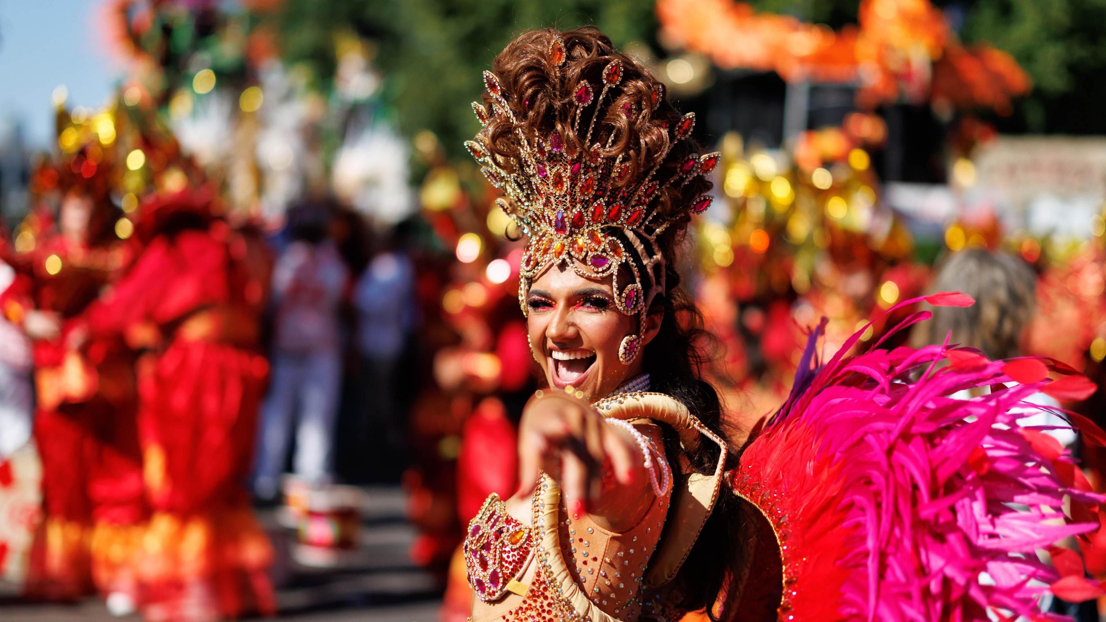 A woman in an orange, pink and red outfit with a beaded headdress points and smiles at the camera. 