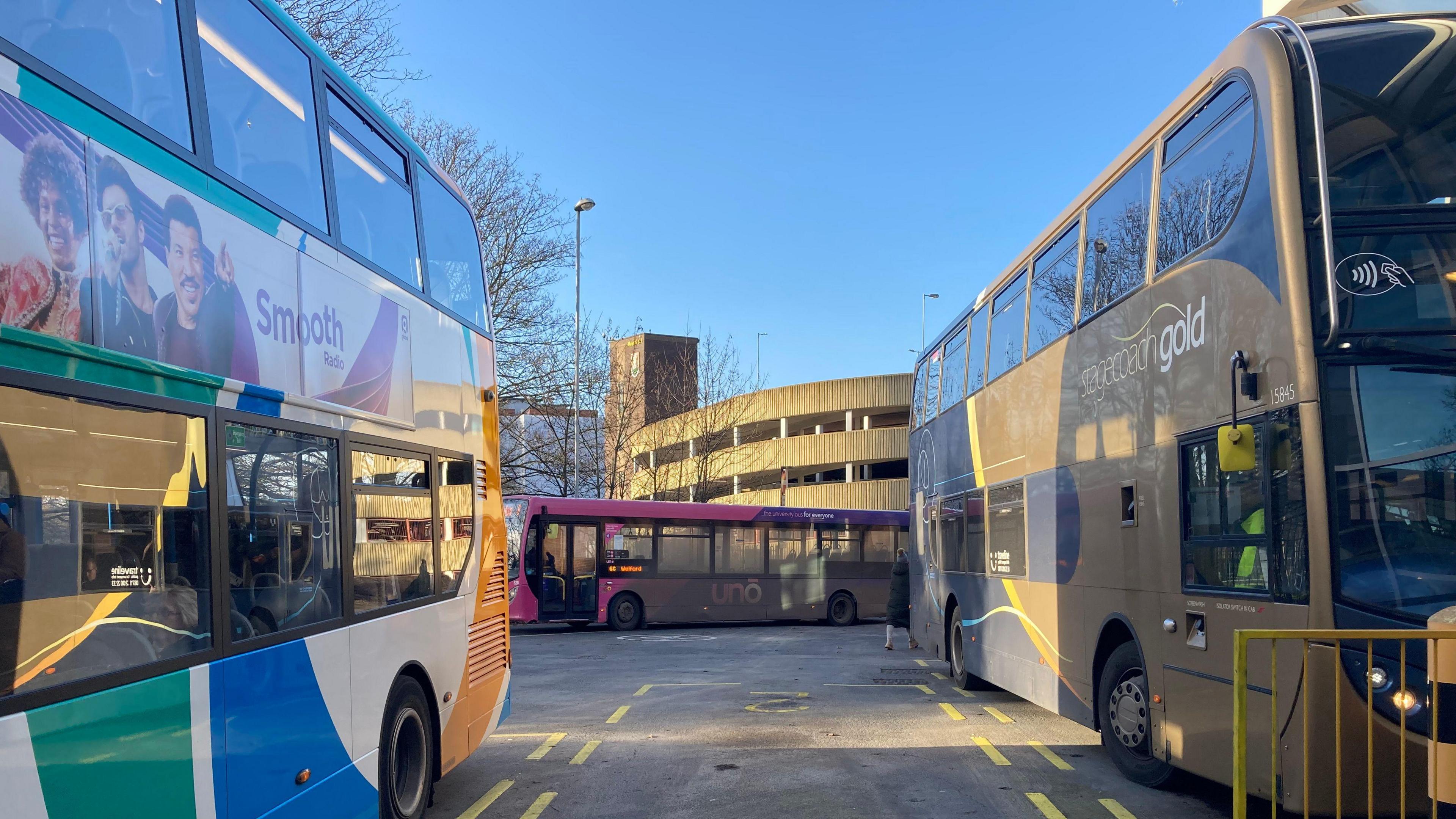 Two double-decker buses parked in yellow numbered spaces in the depot. A single-decker pink bus drives away