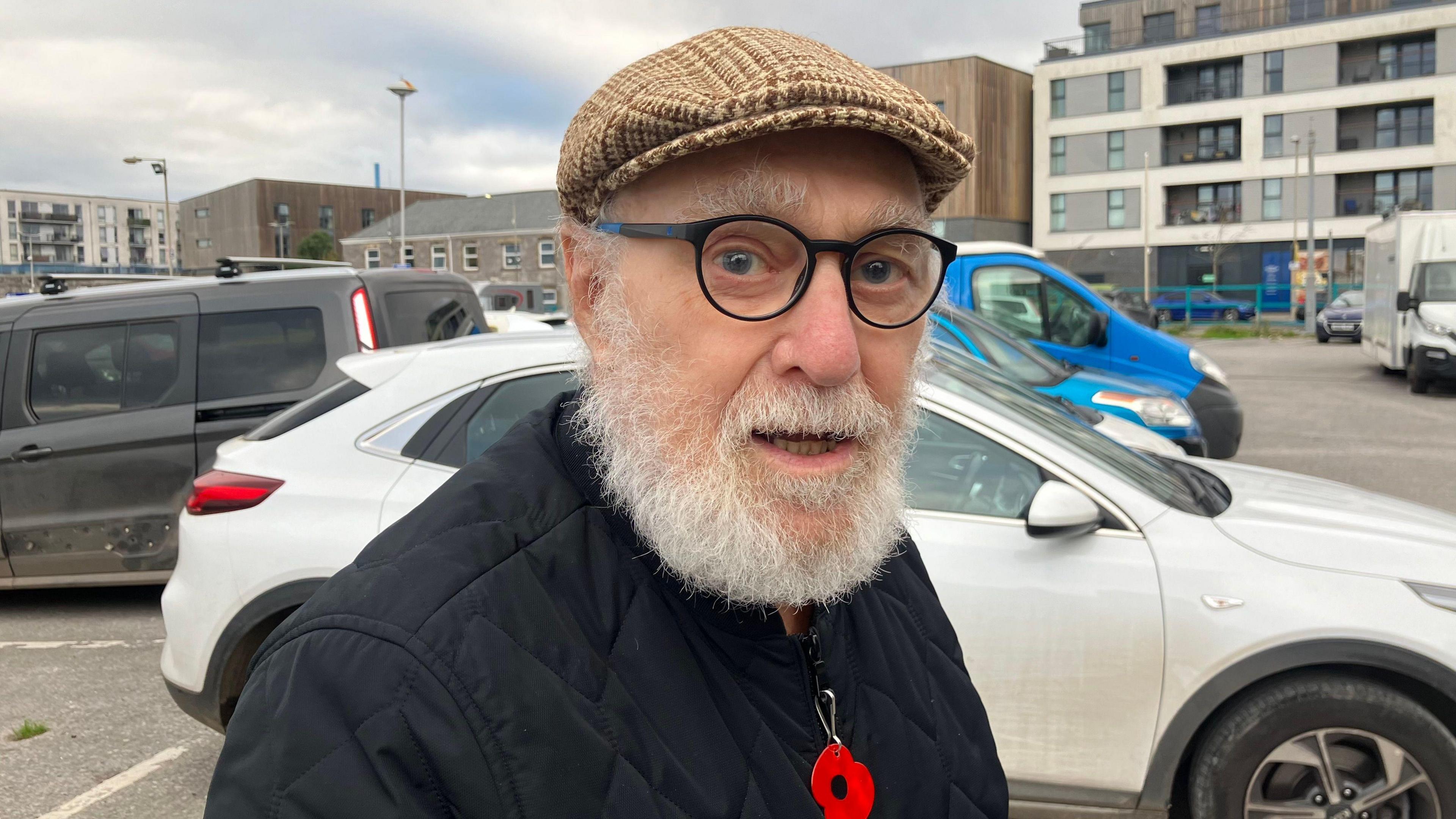 Ian Creasy looking side-on at the camera. He is wearing a brown chequered flat cap with black glasses and a black coat. He has a white beard and is wearing a poppy. In the background is a car park, with a white car behind him.