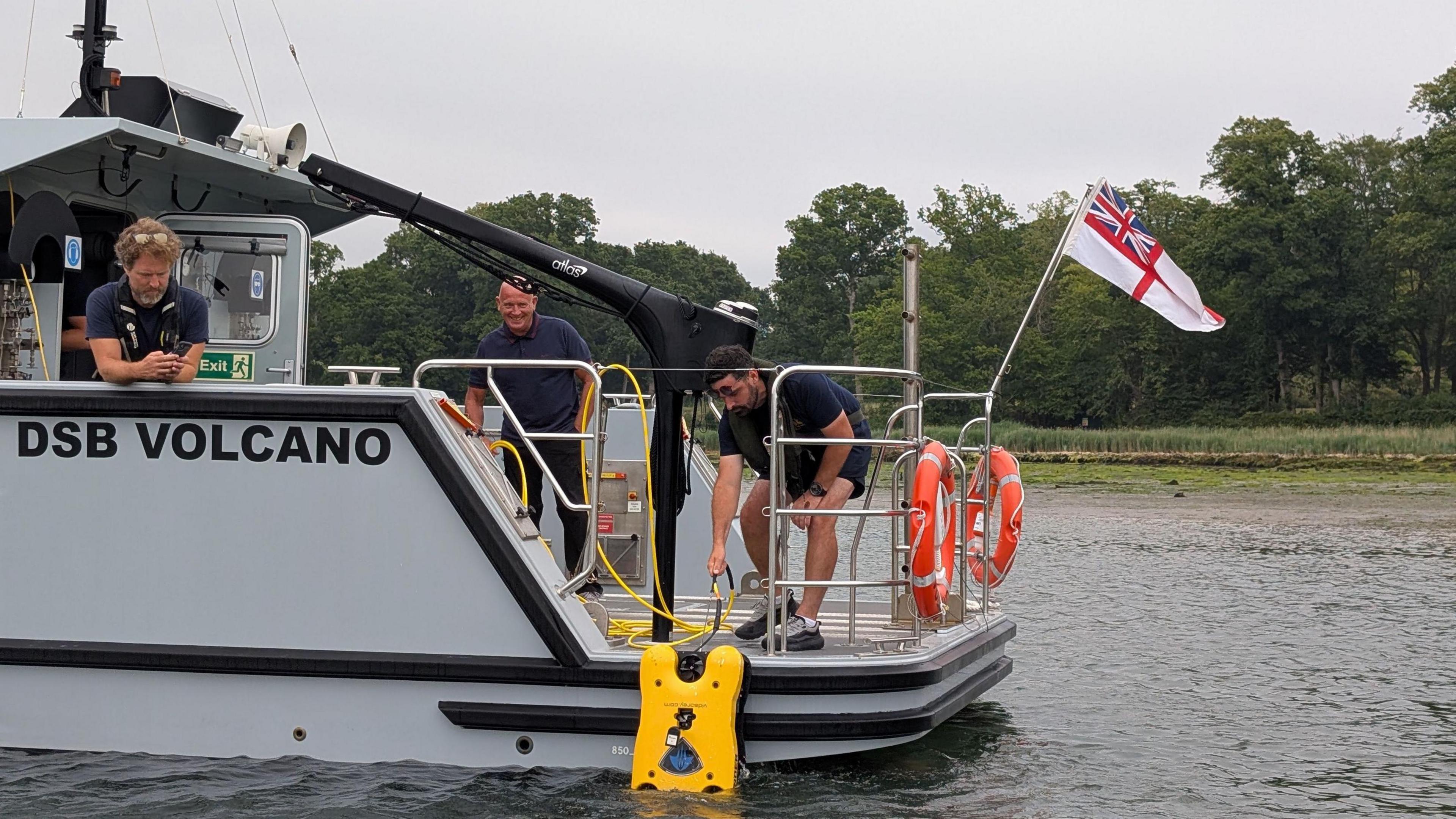 A yellow Remote Operated Vehicle being lowered into the water on a river from the back of a boat where three men are standing. 