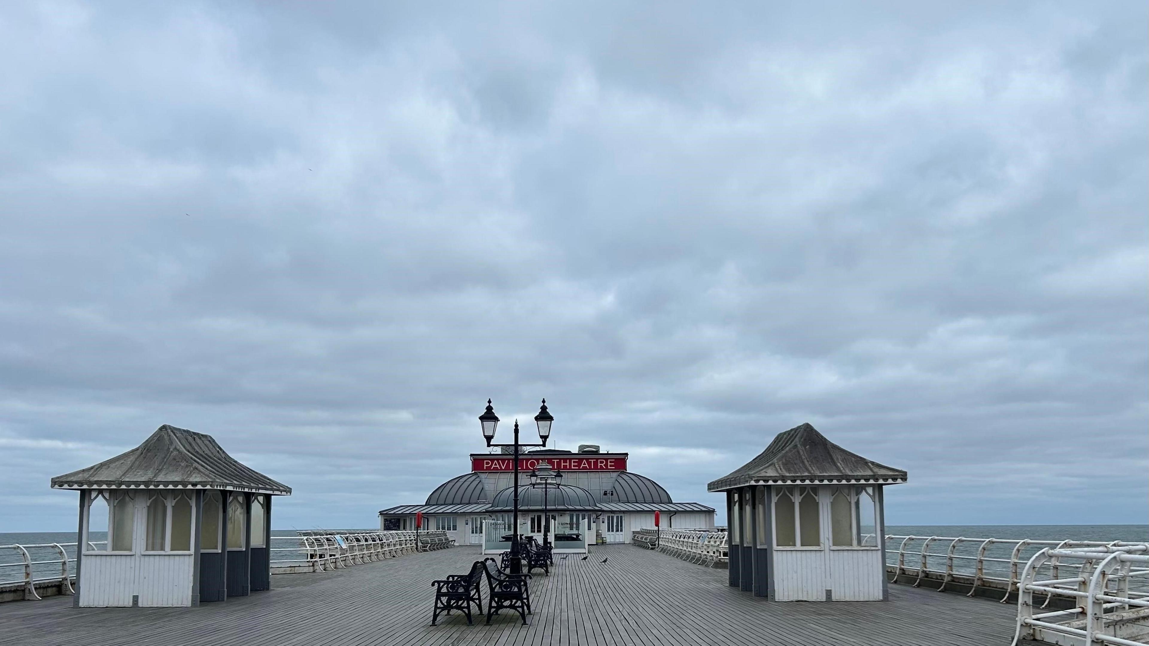 Beige and white wooden pier with two small huts, street lighting, four benches and a pavilion theatre at end of pier. View of sea either side, under a cloudy, great sky