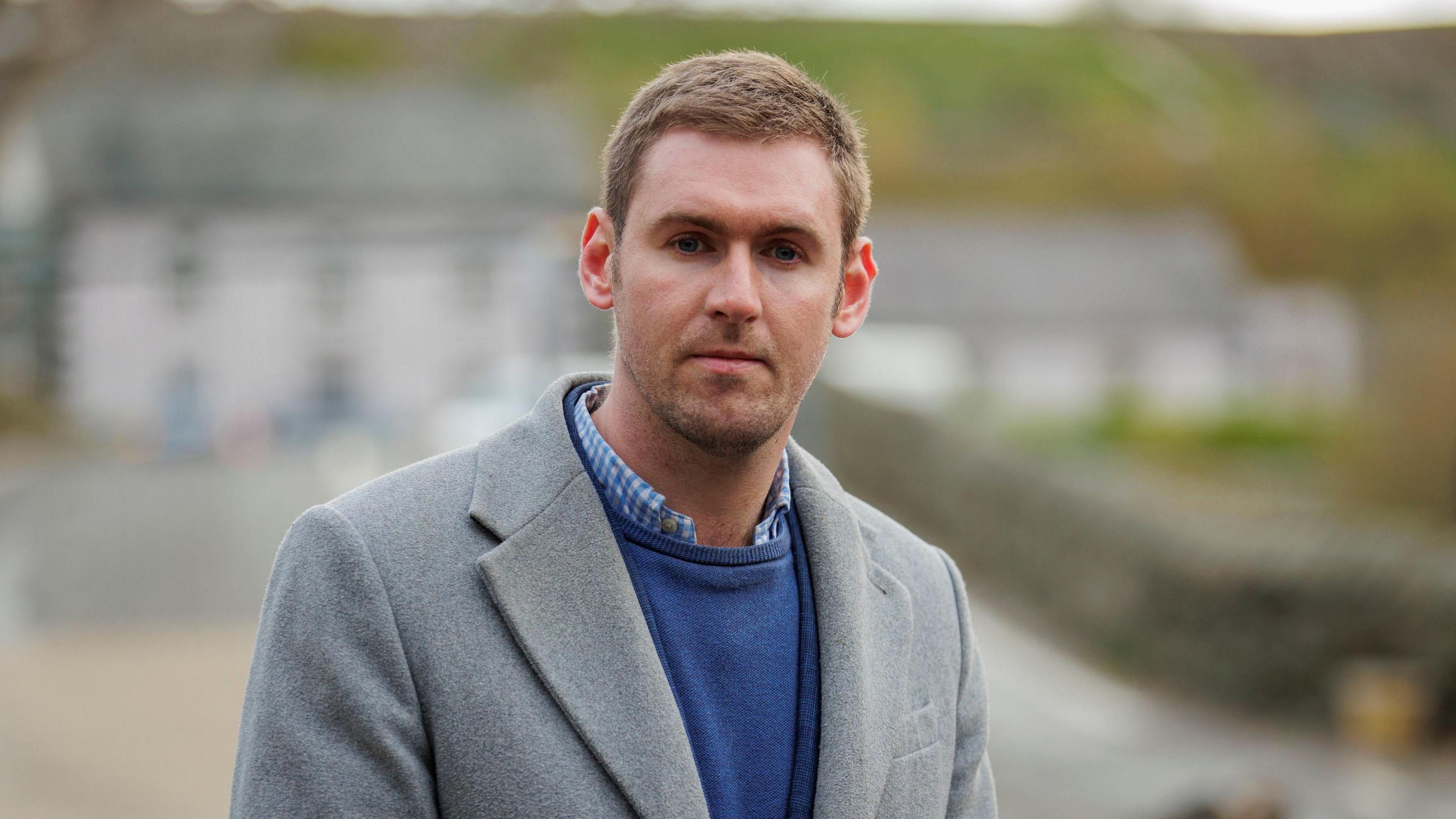 Patrick Brown has short brown hair, and wears a blue jumper, grey jacket and checked shirt. His expression is neutral