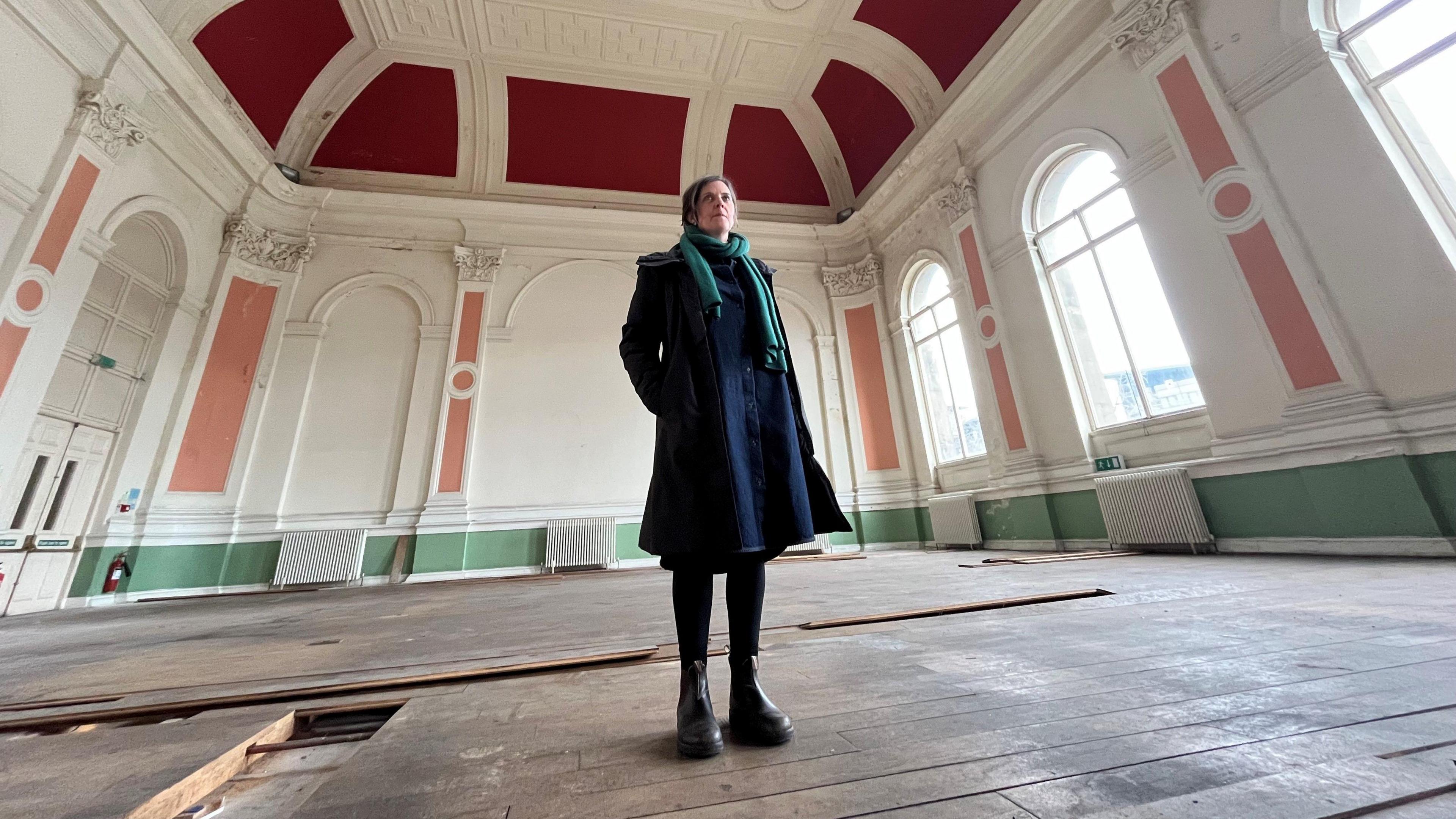 Emily Pieters has short dark hair and is wearing a navy blue smart coat with a teal scarf. She is stood in a large empty room with an ornate ceiling painted in dark red and cream. The room has large windows shaped like arches with peach coloured decorative paint around them.