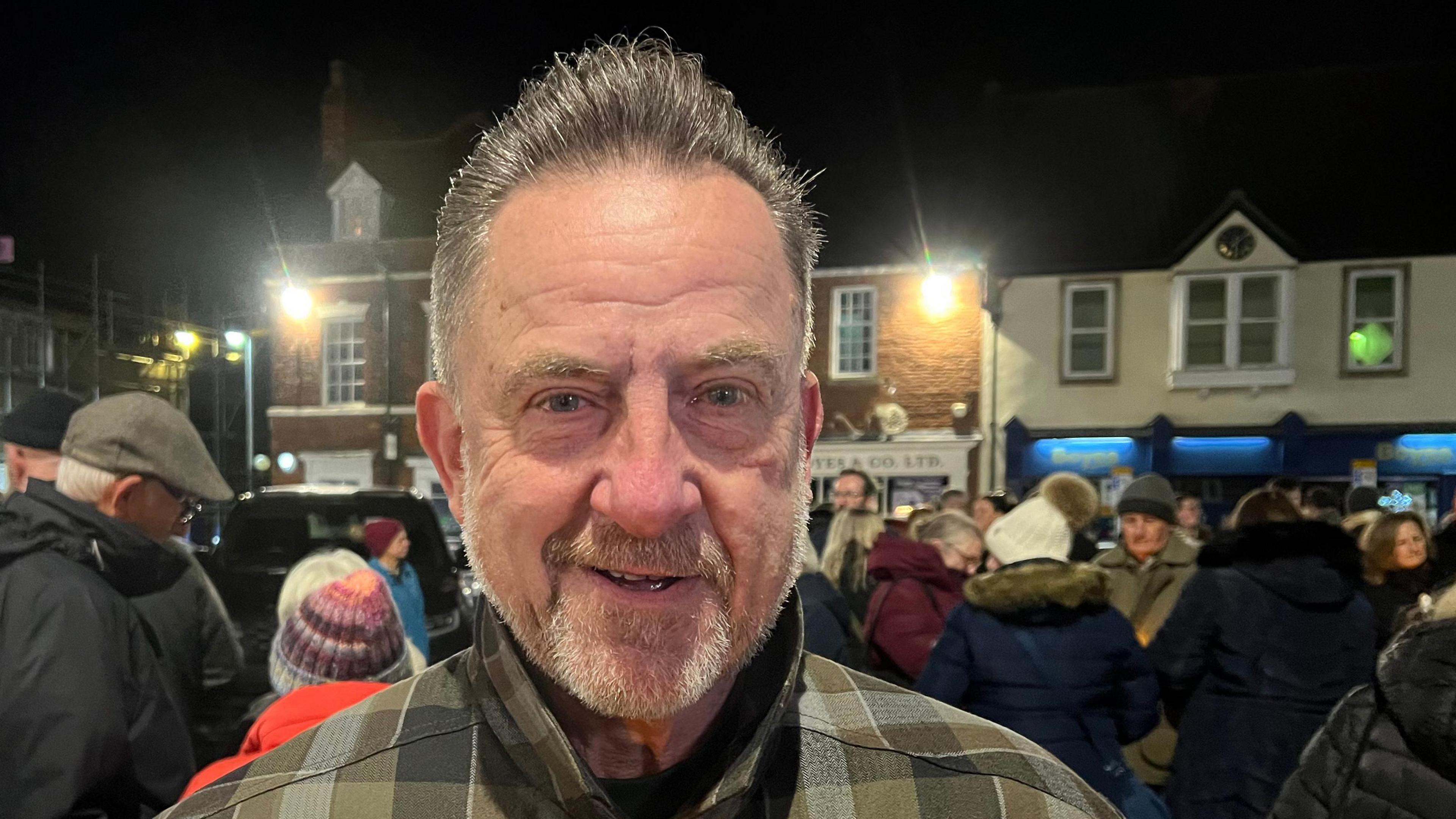 Man with short hair and short beard with chequered jacket. There is a crowd behind him.