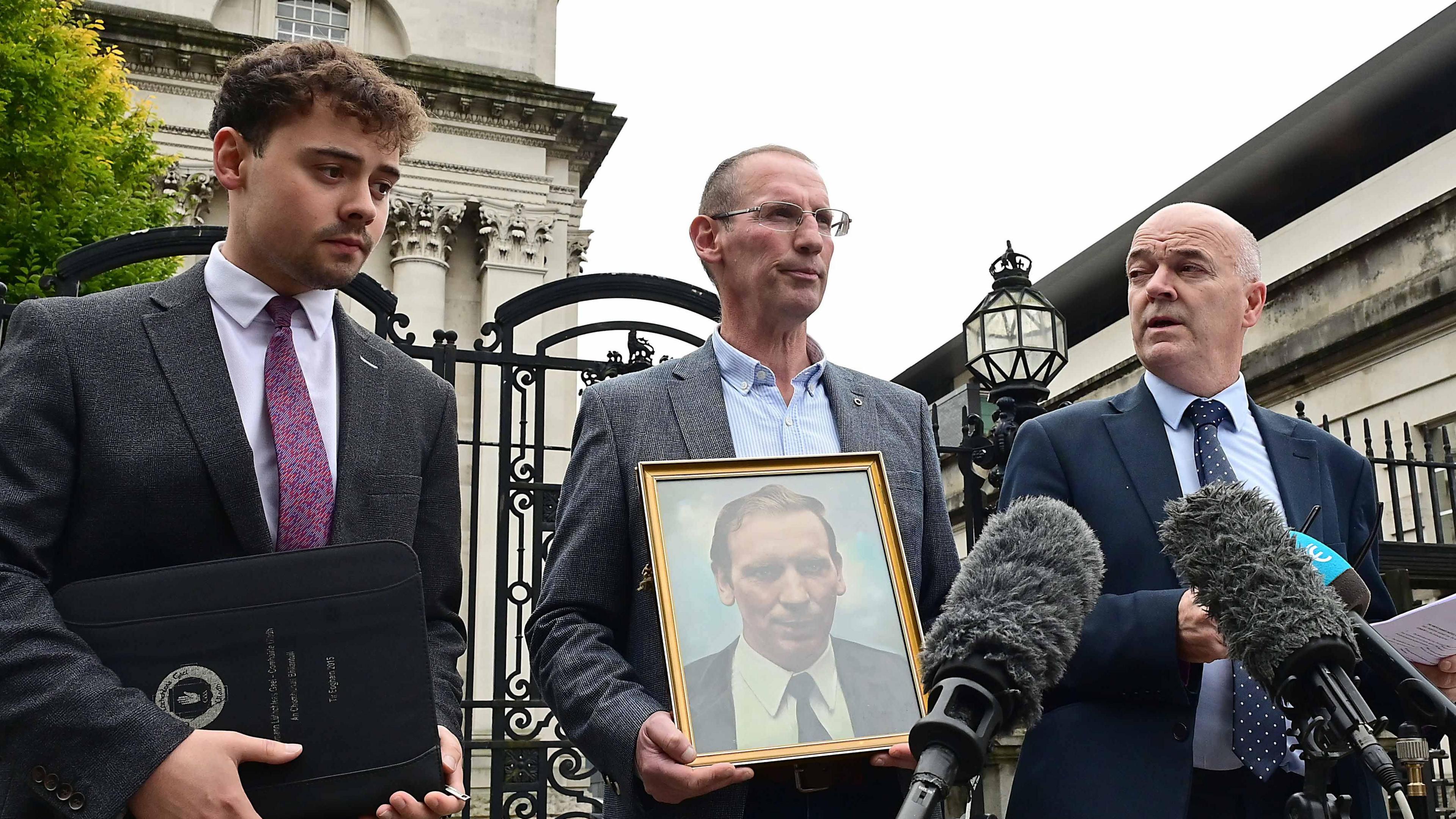 Three men outside court. They wear suits. The man in the centre is holding a gold framed photograph of another man. There are microphones in front of them.