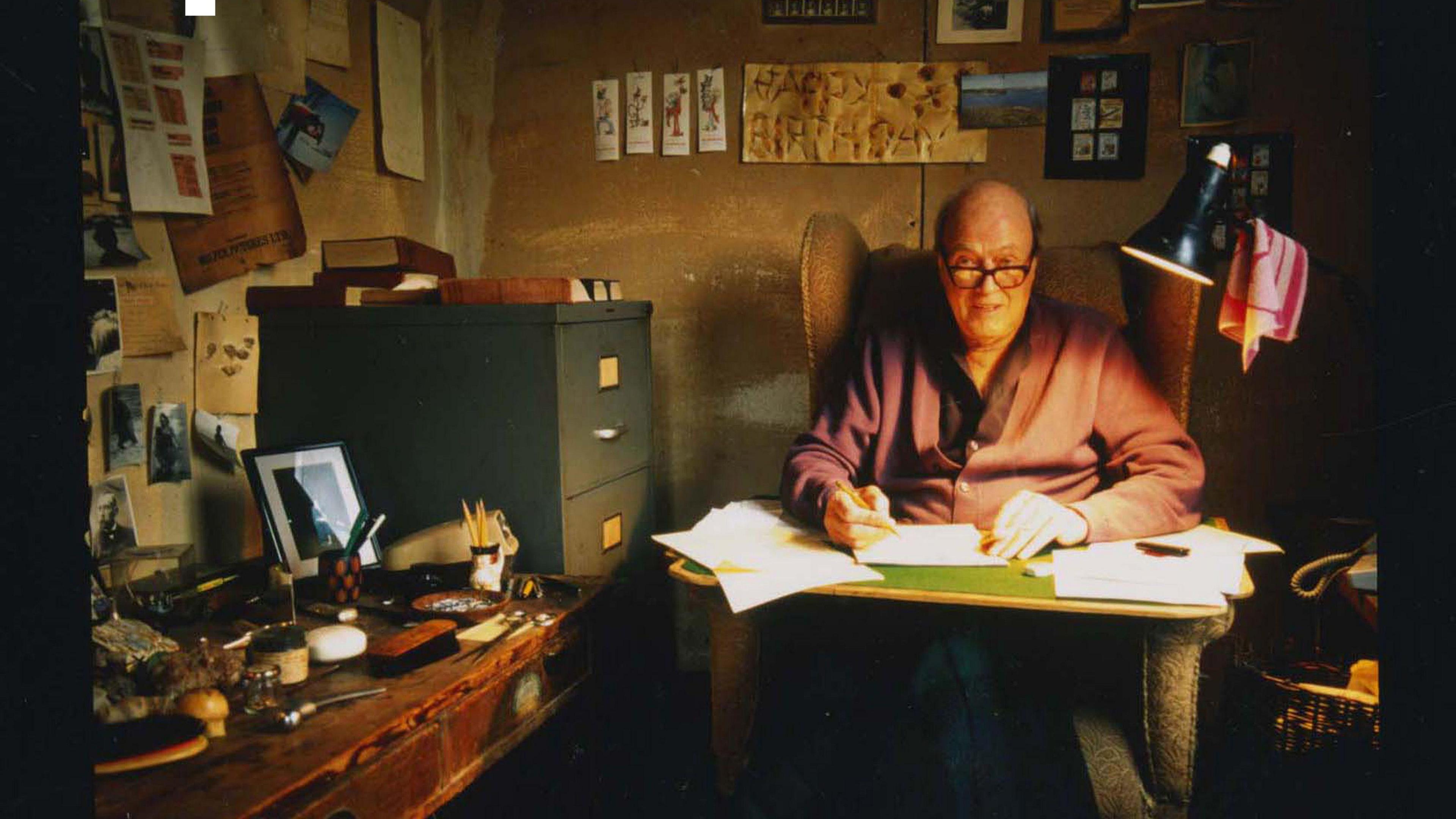 A man with glasses at a writing desk with a lamp over his left shoulder. There is a filing desk to his right and next to it is a desk with lots of different items on it.