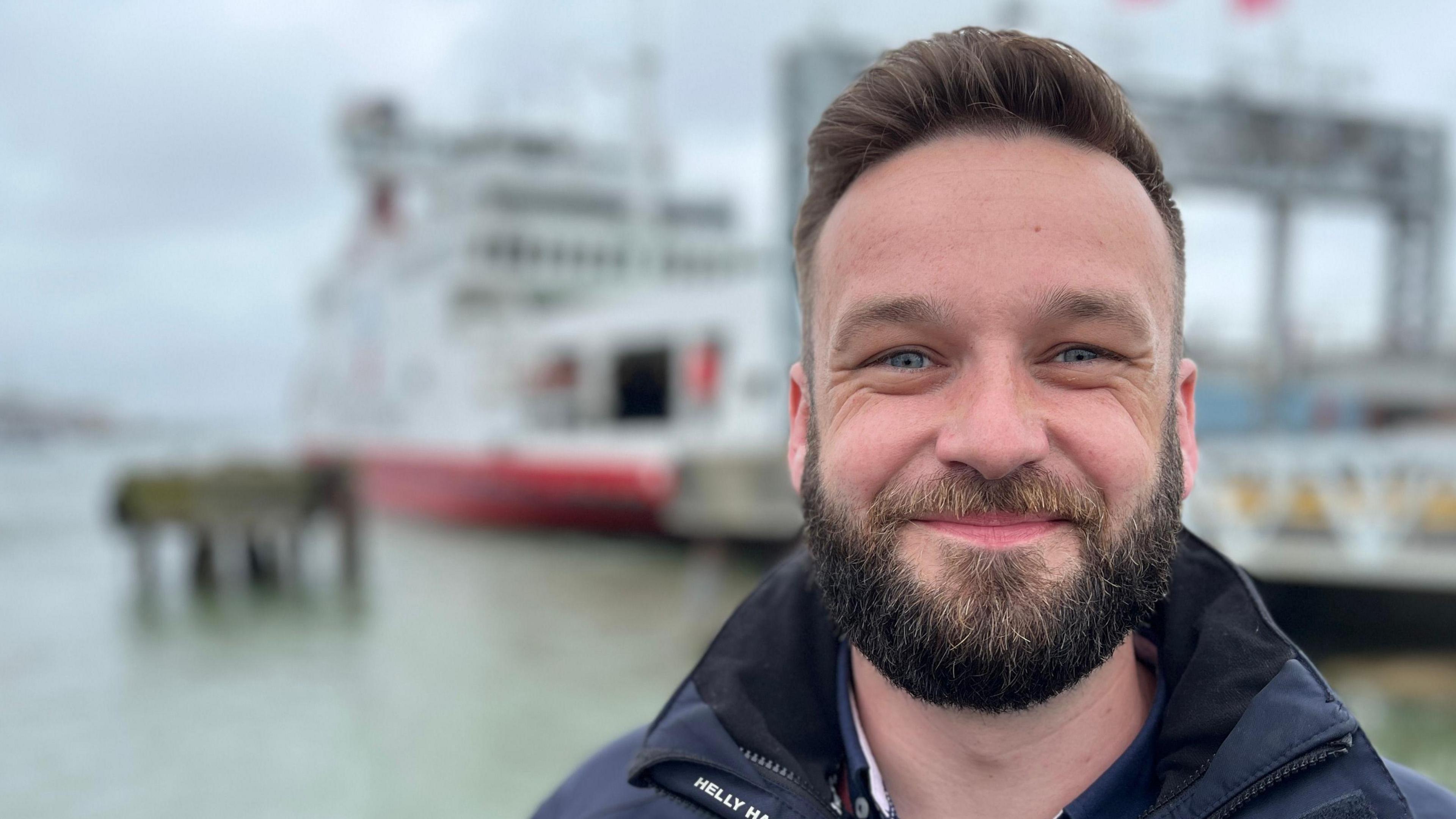 A white man with short brown hair and a brown beard, stood in front of a harbour that is out of focus