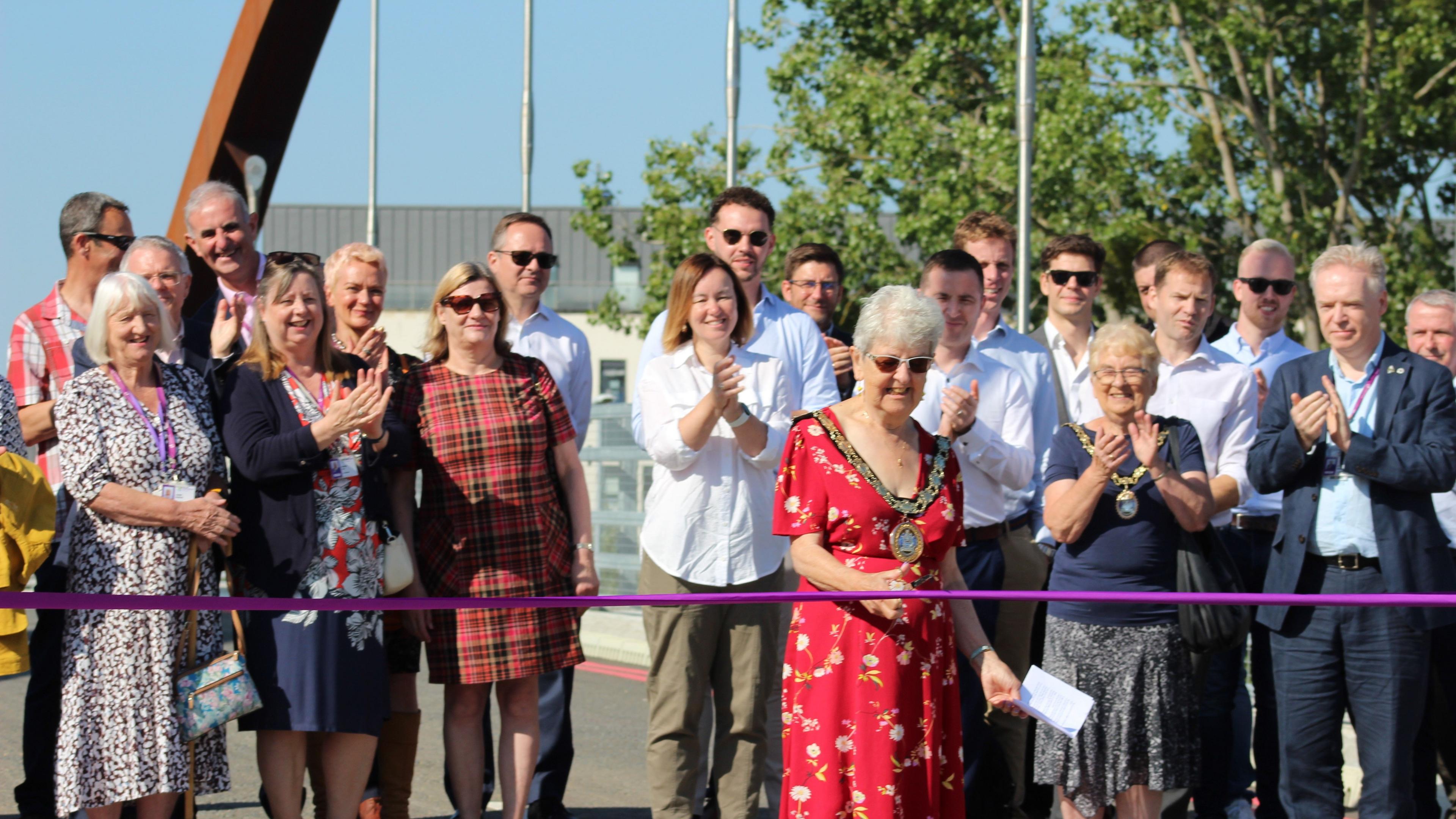 A group of spectators applauding as a ribbon is cut by a woman in a red dress and a mayoral collar.