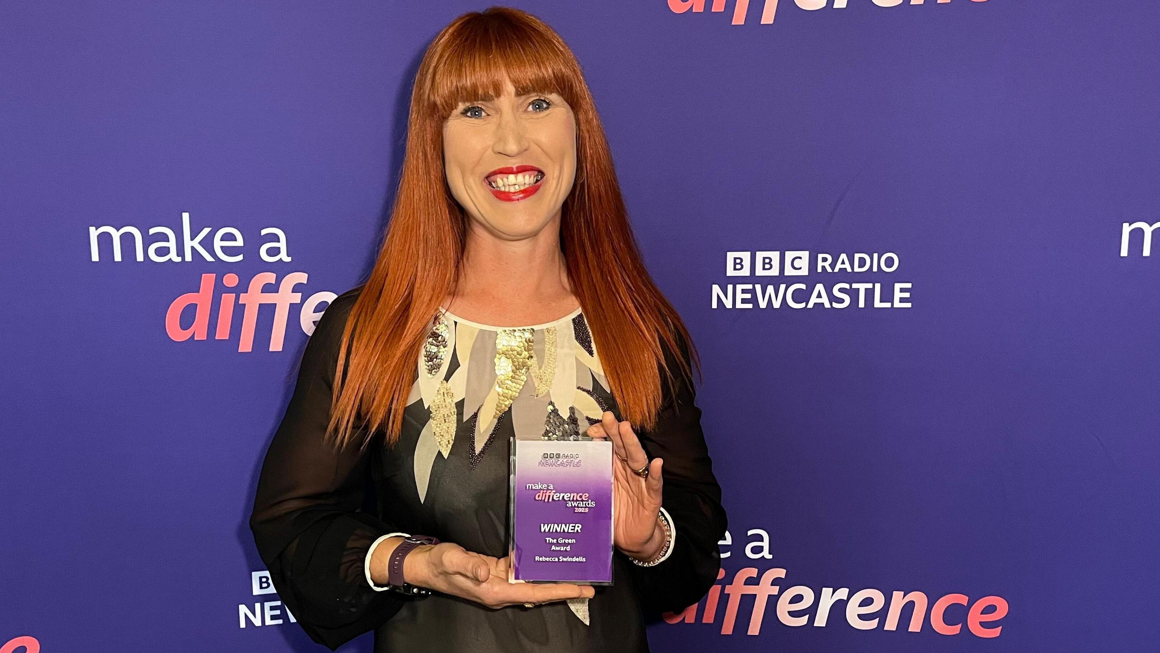 Rebecca Swindells is standing with an award in front of purple signage that reads 'MAKE A DIFFERENCE' with the BBC Radio Newcastle branding. She has long red hair and wears bright red lipstick and a black dress with metallic patterns on the shoulders.