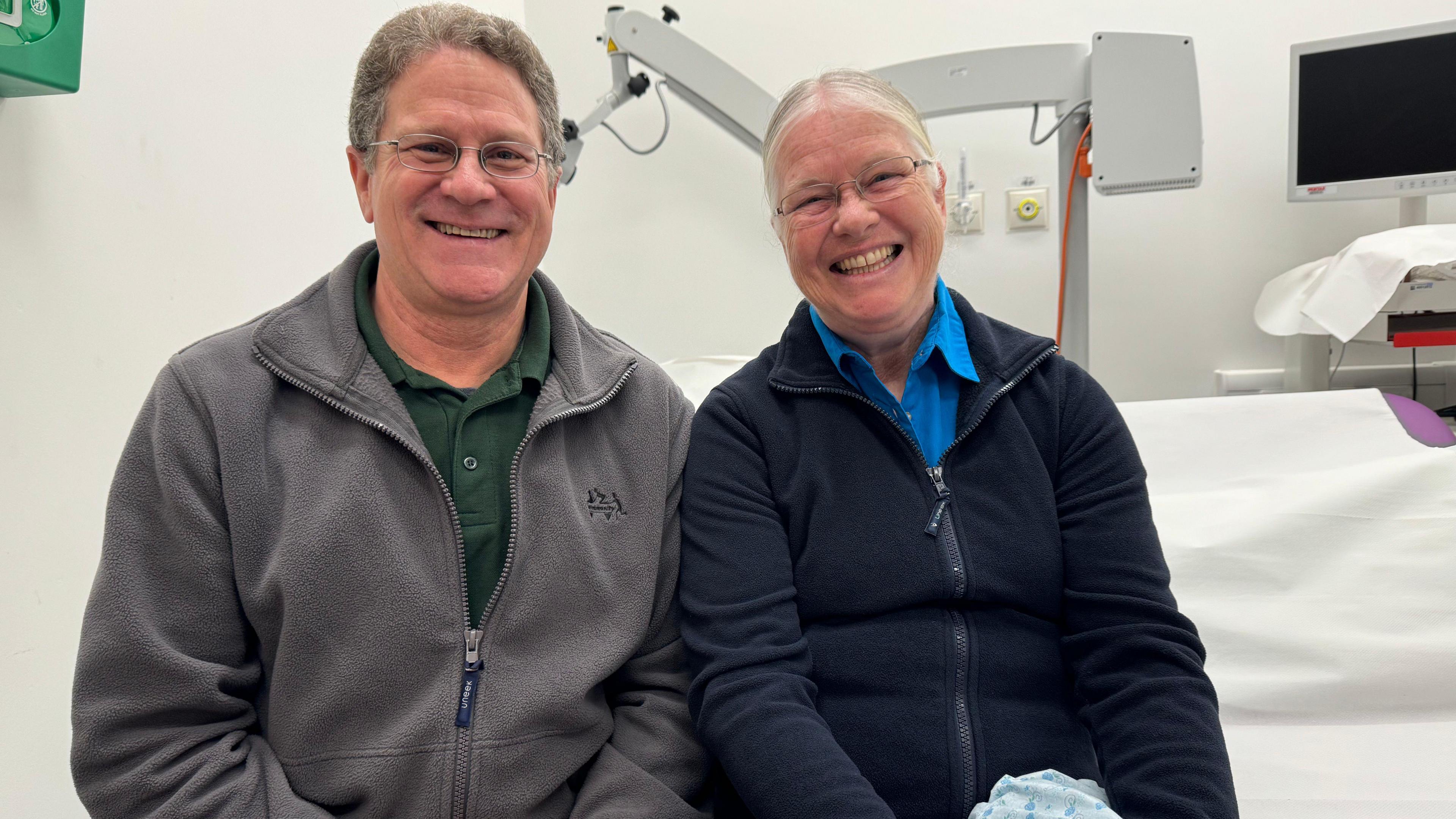 Kim Boller, a man in his 60s sits next to his wife Natalie, also in her 60s. Kim wears a zip up grey sweater and green shirt and Natalie wears a light blue shirt and dark blue zip up. Both are smiling.