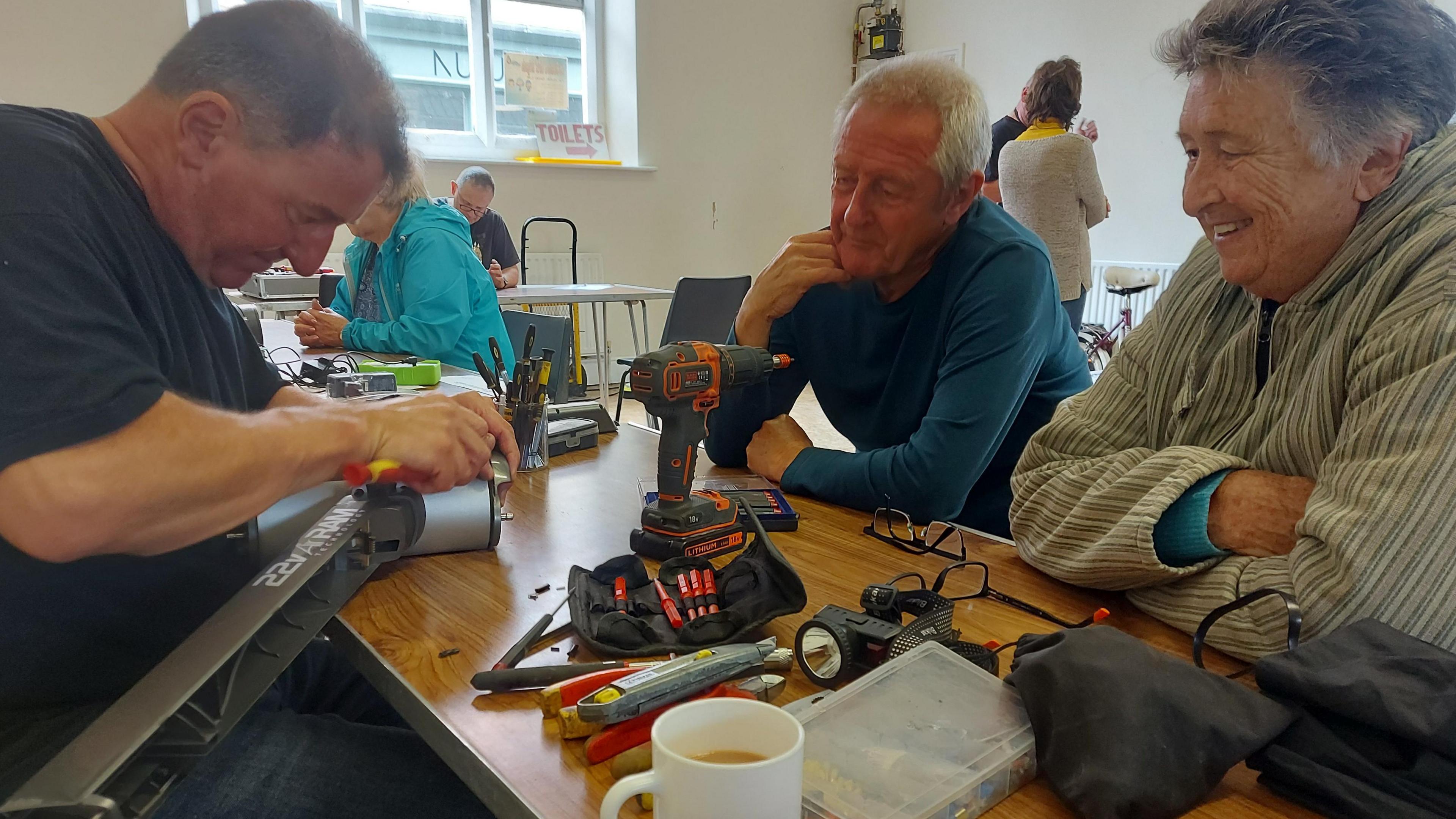 A man is mending an item and concentrating as a couple sitting opposite him watch him work. They are smiling. They are sitting at a table in a church hall and there are tools scattered on top of the table. This is a repair cafe where people can bring broken items to be repaired. 