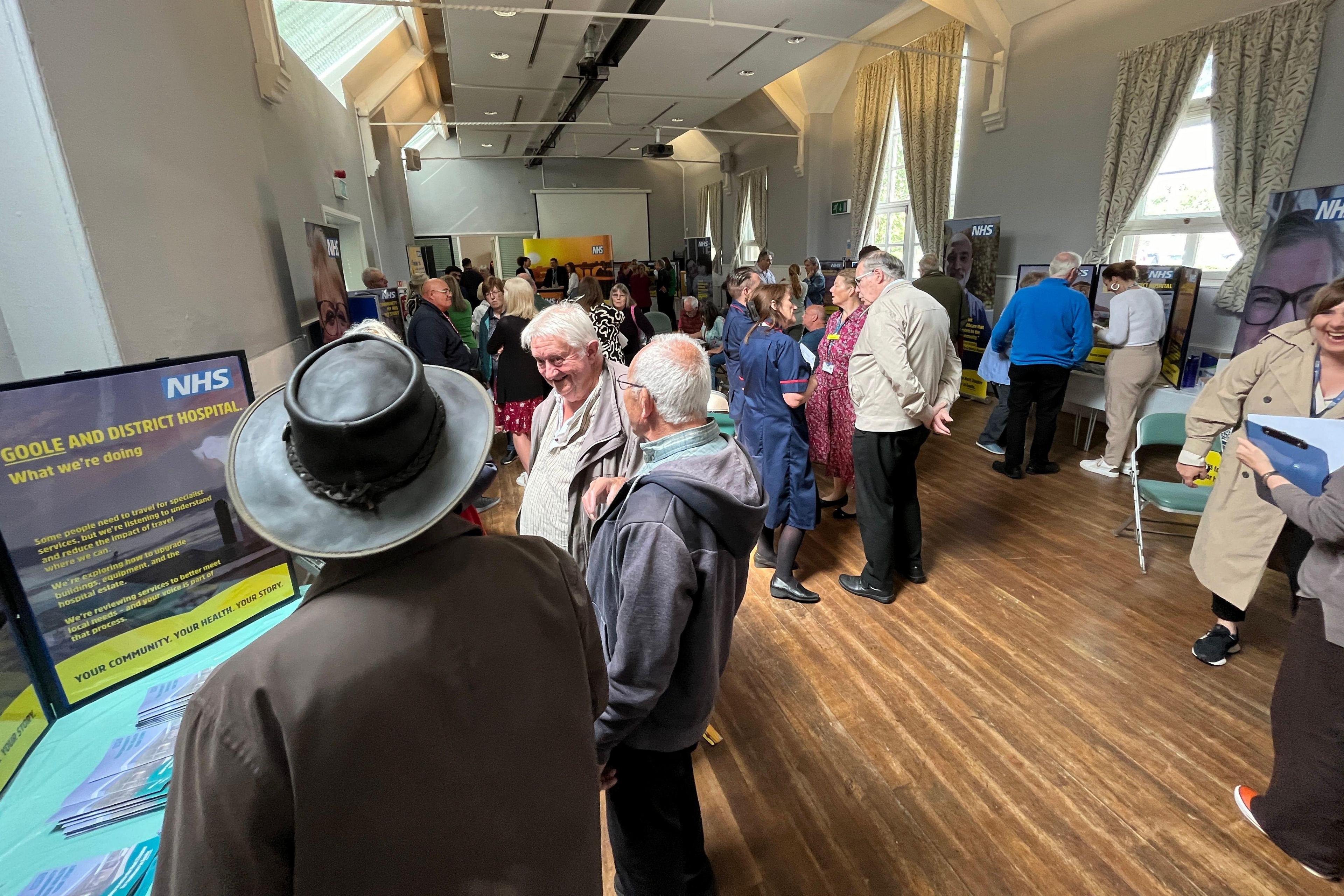 People standing in a room at a drop-in session. There is a wooden floor and a display on a table with "NHS" written on it.
