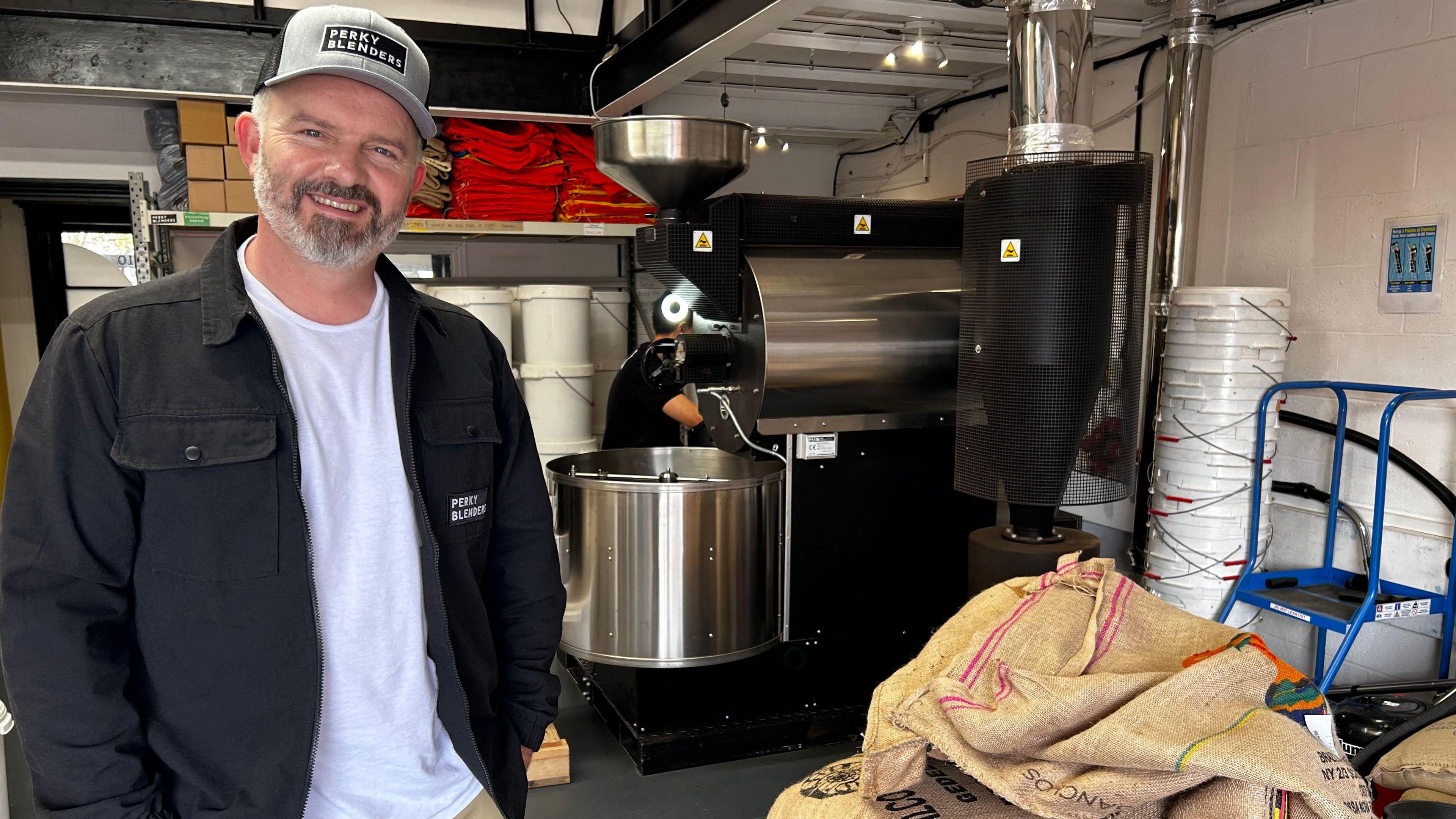 A man called Adam standing in front of a large, commercial coffee roasting machine. He has a beard and is wearing a grey cap and black jacket that say Perky Blenders.