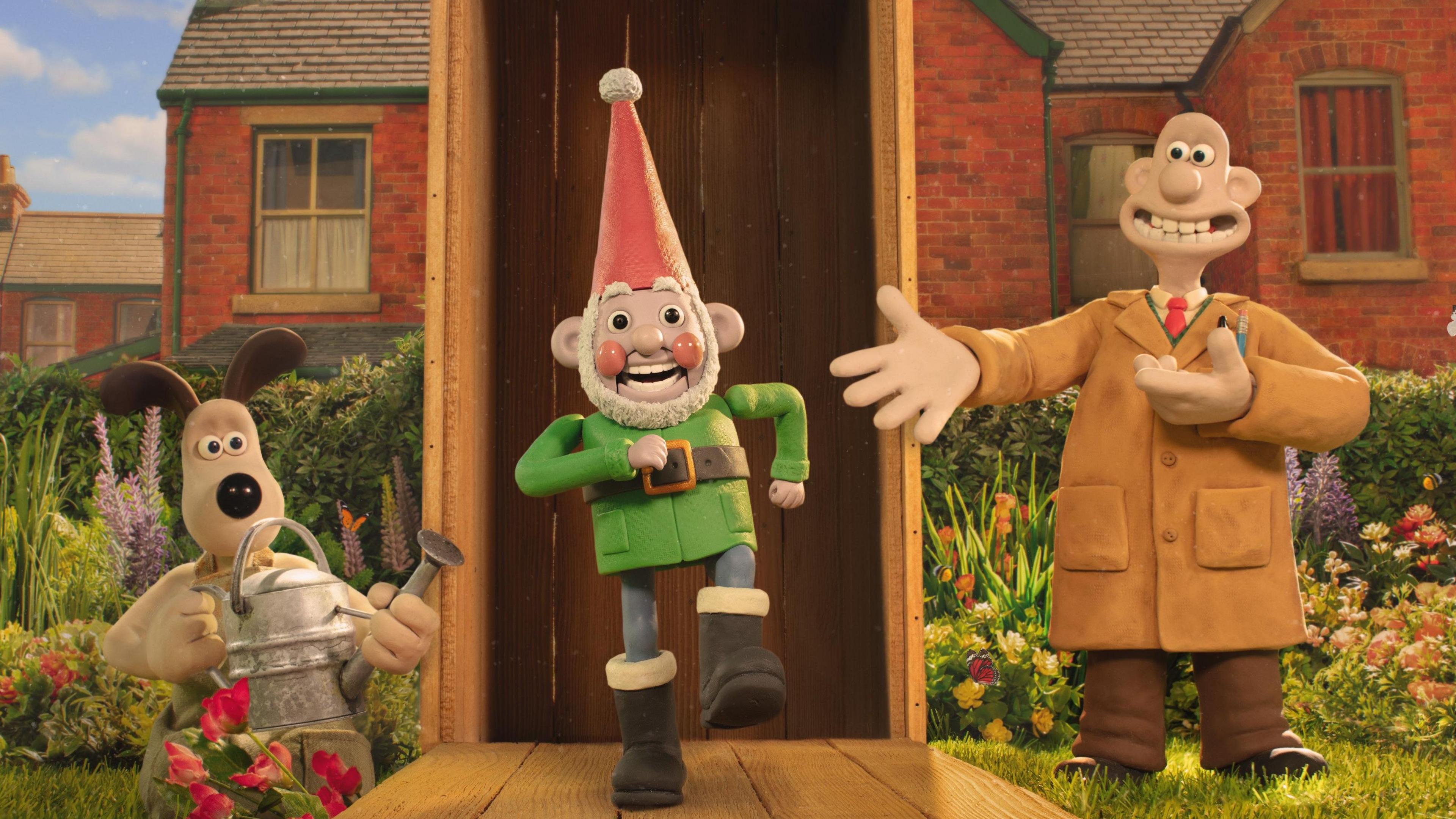 An animated dog with ears sticking up holds a watering can and peers out of a flower border at a garden gnome which is grinning and in a marching stance. On the other side of the gnome stands an animated man in a brown overall with a big wide toothy smile, standing on a lawn with flower beds behind him and the rear of red brick terraced houses in the background. 