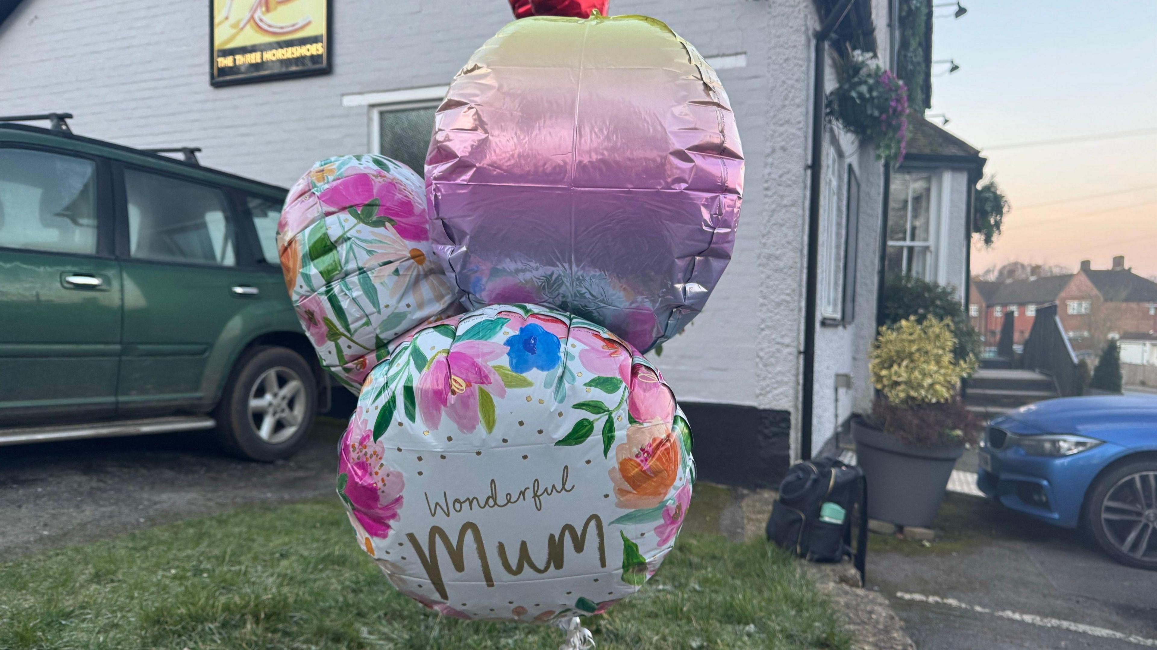 Three balloons, including one saying 'Wonderful Mum', are tethered to the ground in front of the Three Horseshoes pub in Knockholt. A green and a blue car are parked behind the balloons.