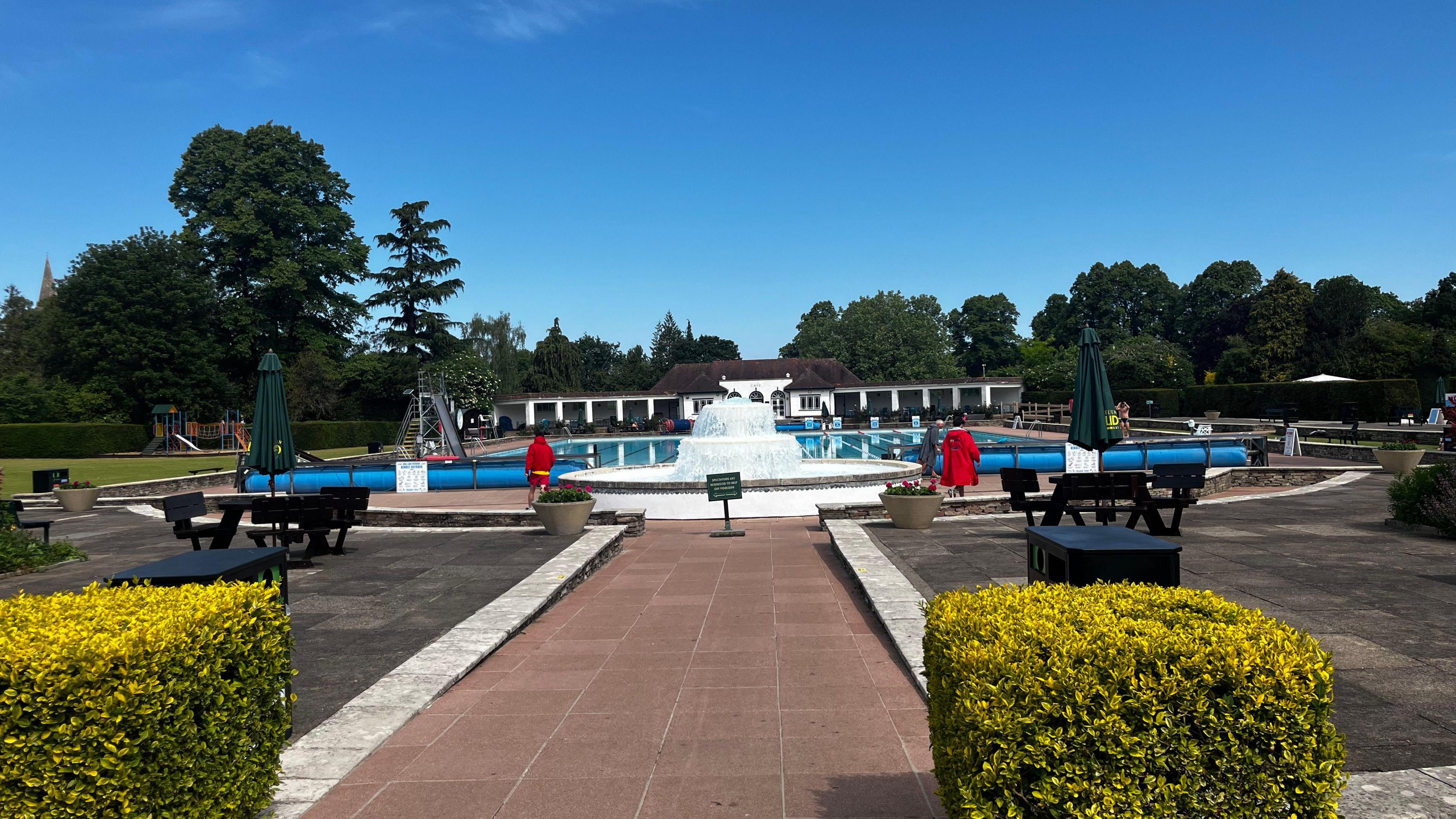 Image shows Cheltenham's Lido with a blue sky in the background. Two lifeguards can be scene walking around the pool in red clothing.