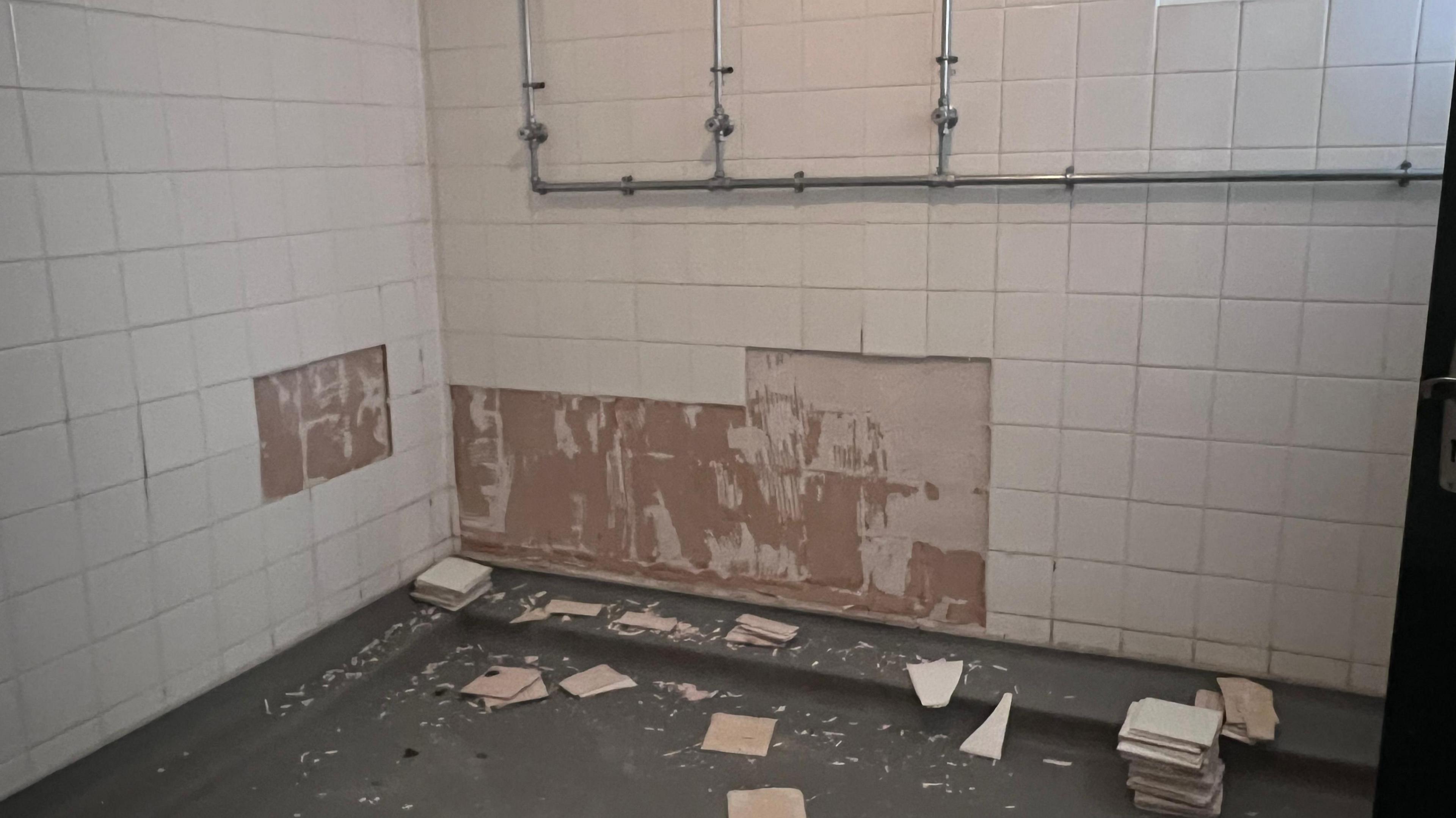 Picture shows a wall with three showers missing many tiles. There is a lot of broken tiles on the ground and a pile of new ones.