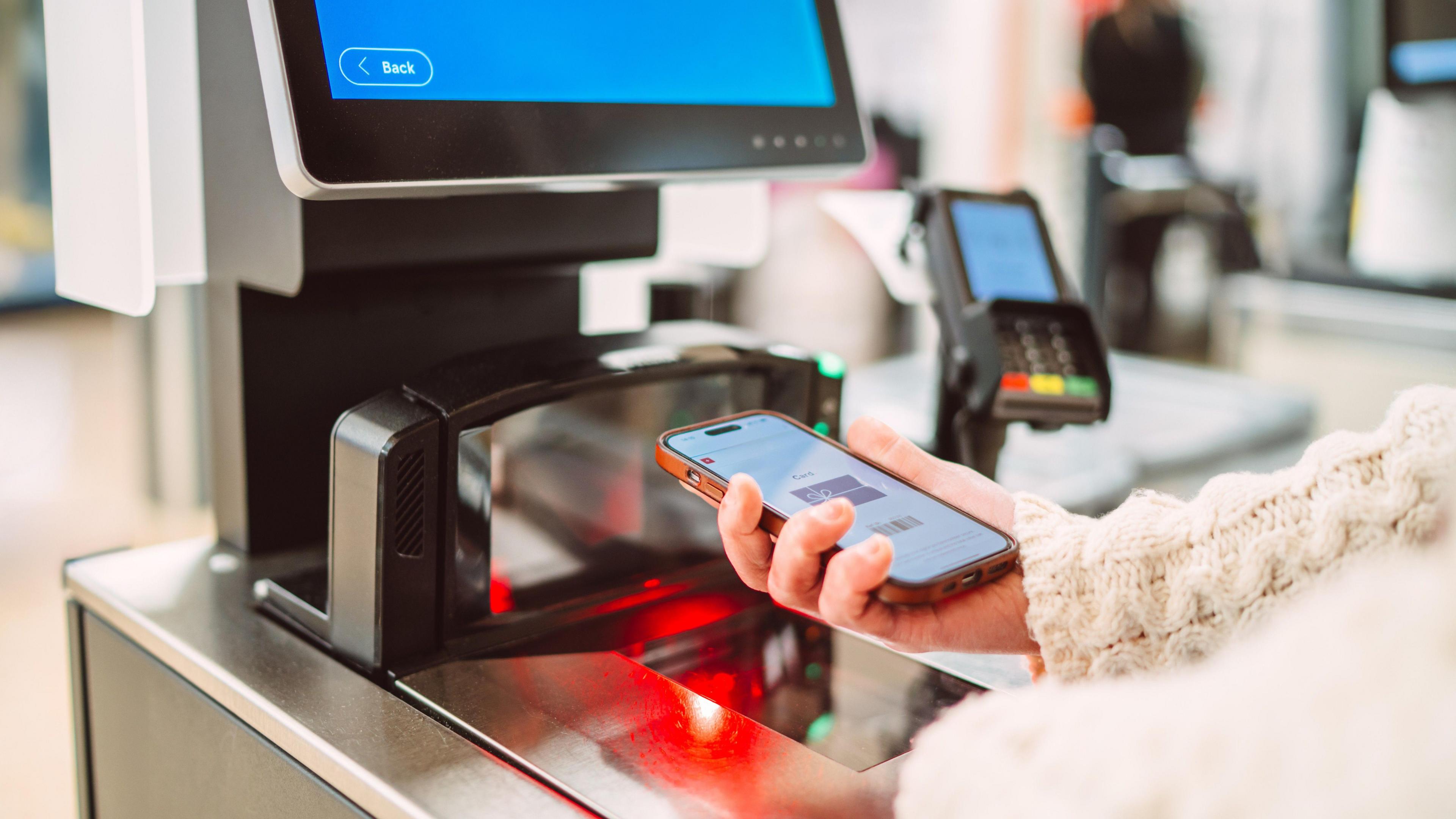 Close-up of a shopper using a smart phone to scan a digital barcode at a self-checkout terminal in a modern retail store.