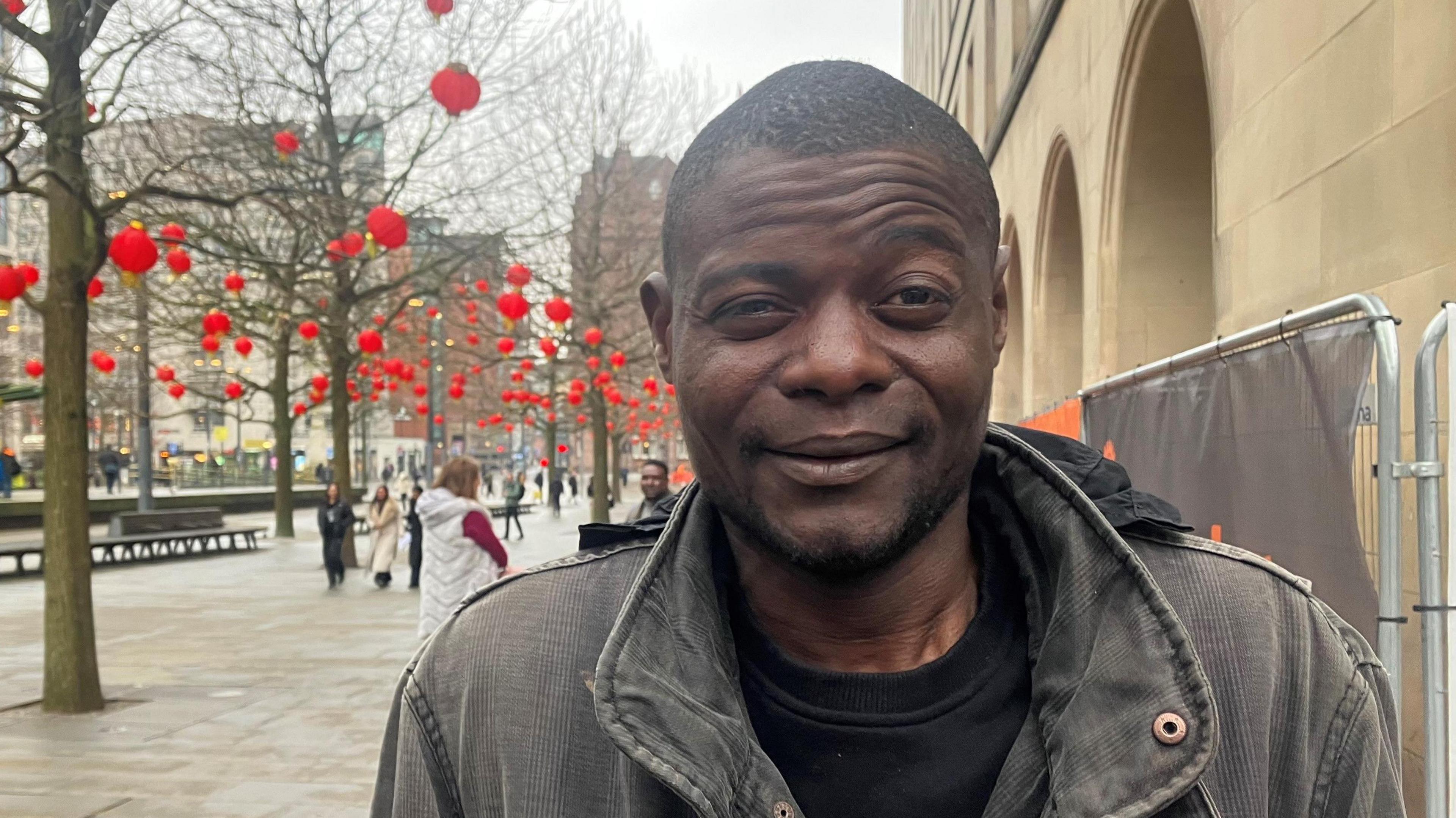 Mamadi Camara from Equatorial Guinea wearing a grey jacket and black t-shirt stands beneath the archways of St Peter's Square on an overcast day. Trees with Chinese lanterns hanging from them can be seen behind him as people mill about the square.