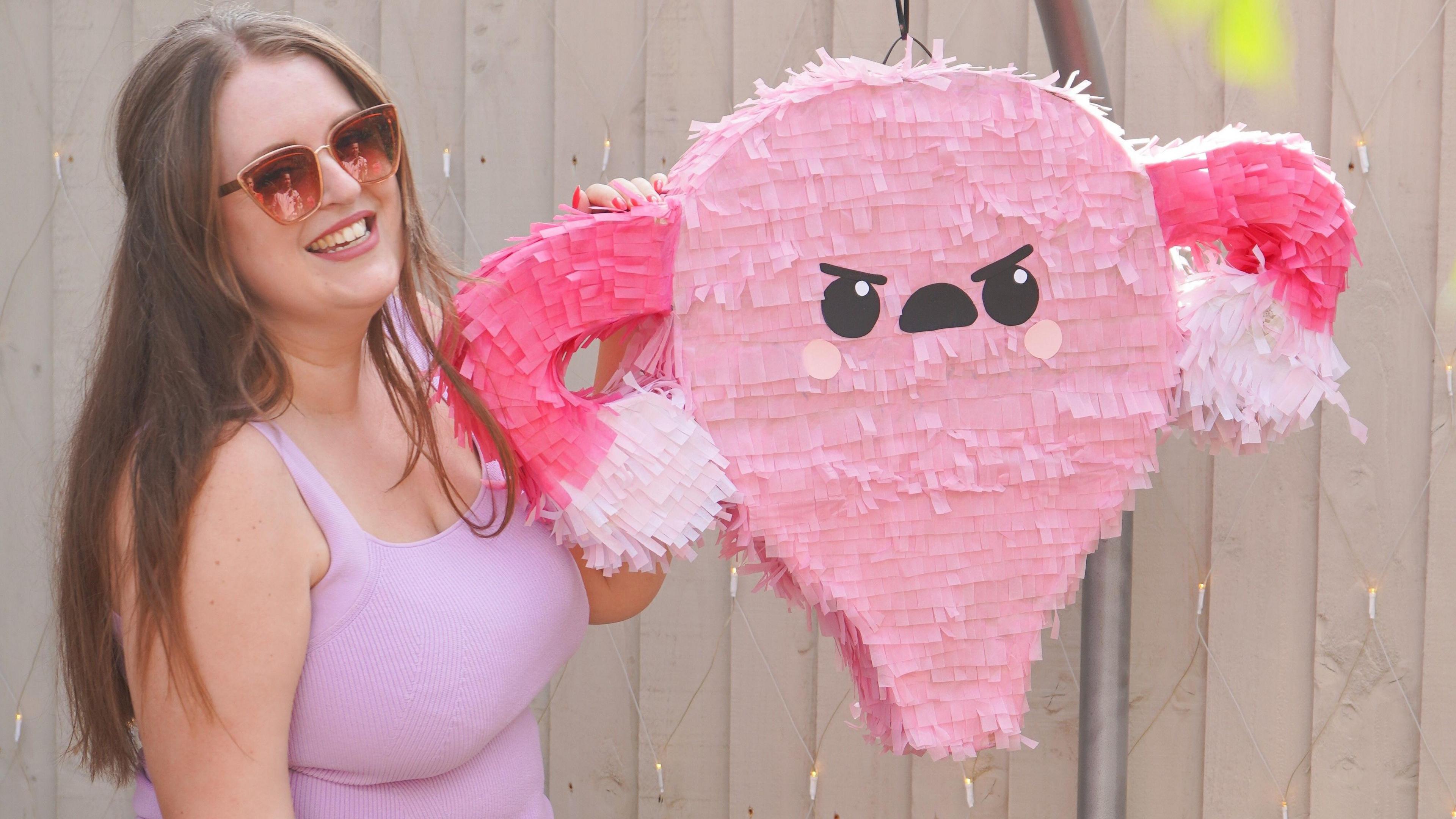 Jen is wearing a pink top and sunglasses, she is standing next to a pink pinata in the shape of a womb and ovaries.