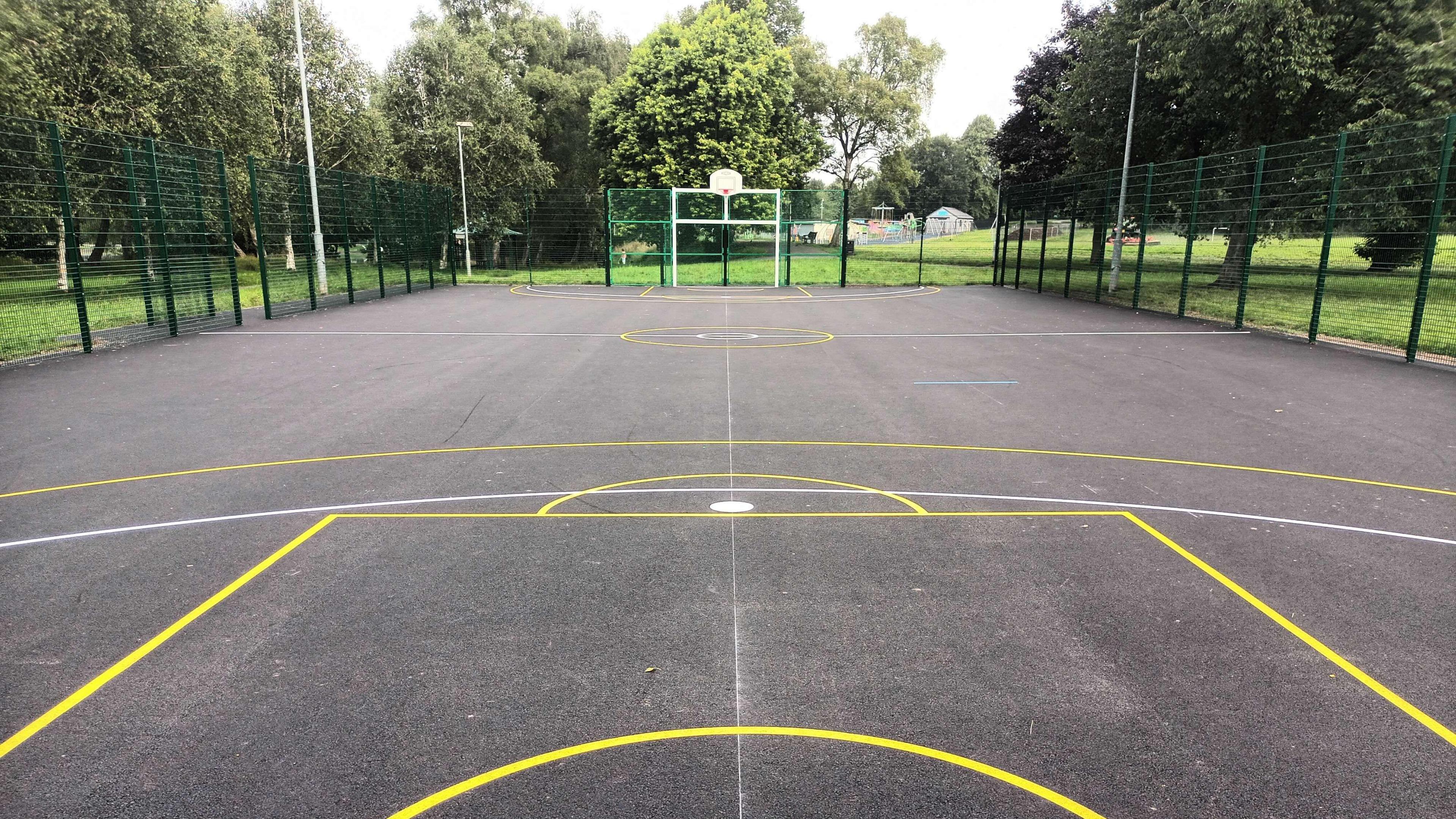 The refurbished multi use games area. The playing pitch is within the park, surrounded by grass and trees. It has goal posts and basket ball hoops and lines of different colours on the ground to show the pitch areas for different games.