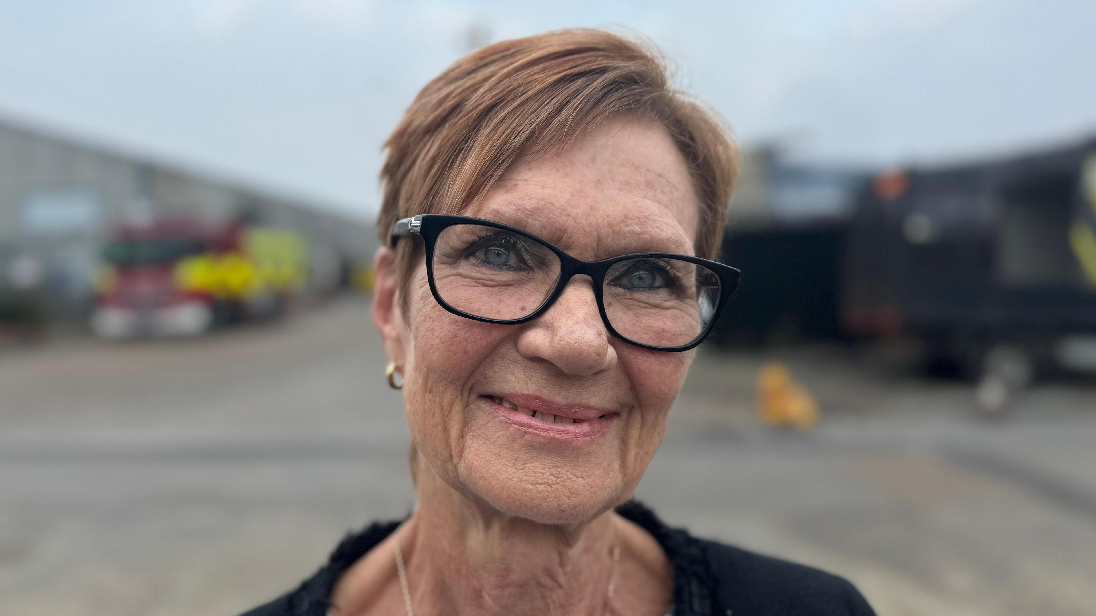 A smiling woman wearing glasses. A fire engine is in the background.