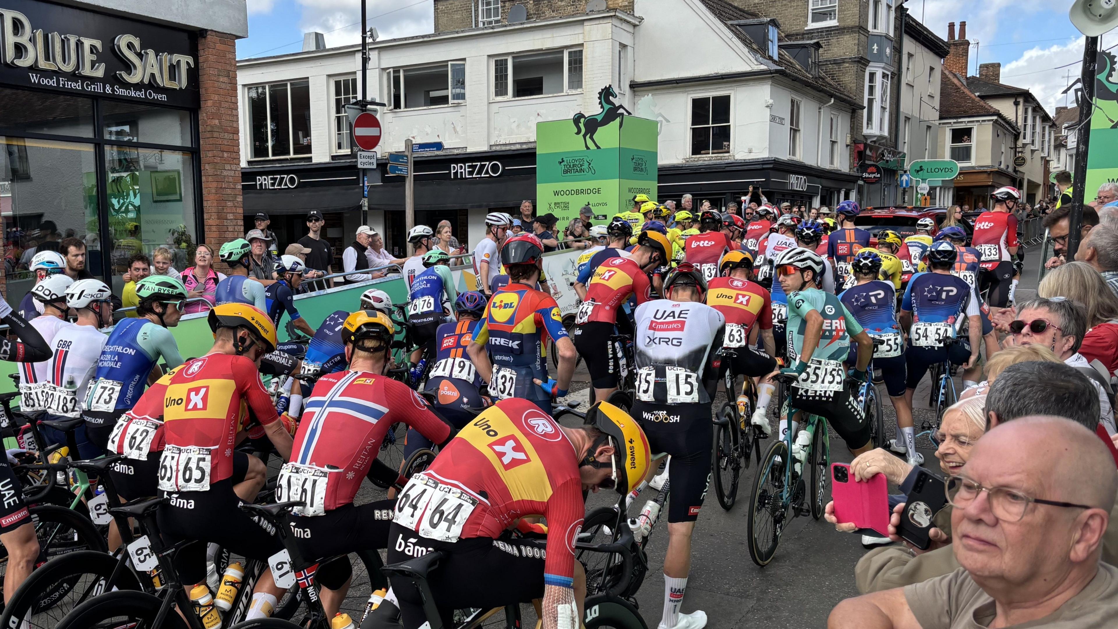 Tens of cyclists sit at the start of the Tour of Britain Men race waiting to get going. Crowds of people watch on from behind railings either side of the road.