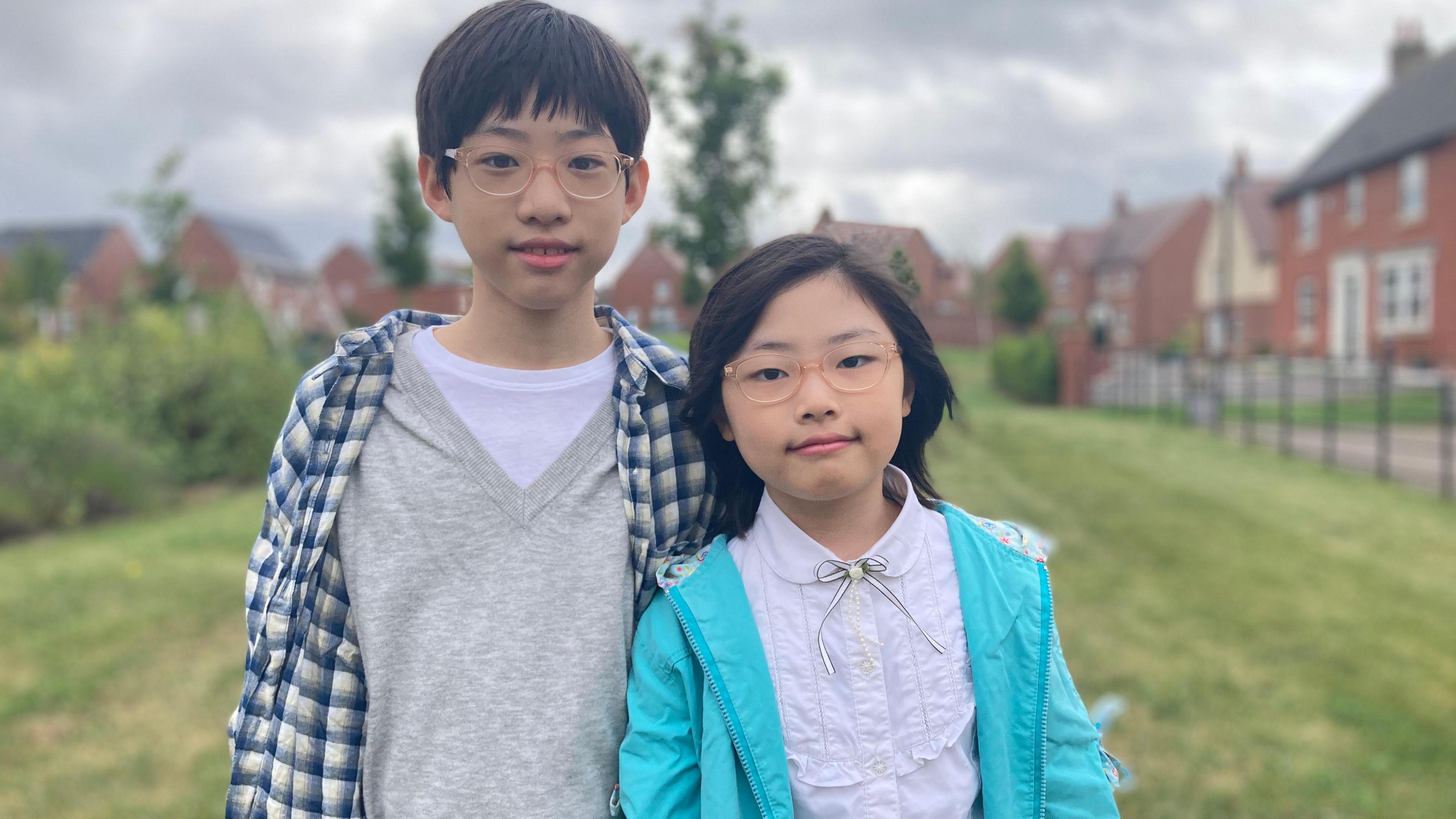 Elias, a boy wearing a grey and white shirt with a blue checked overshirt, standing next to his sister Ariel, wearing a white shirt and light blue jacket.