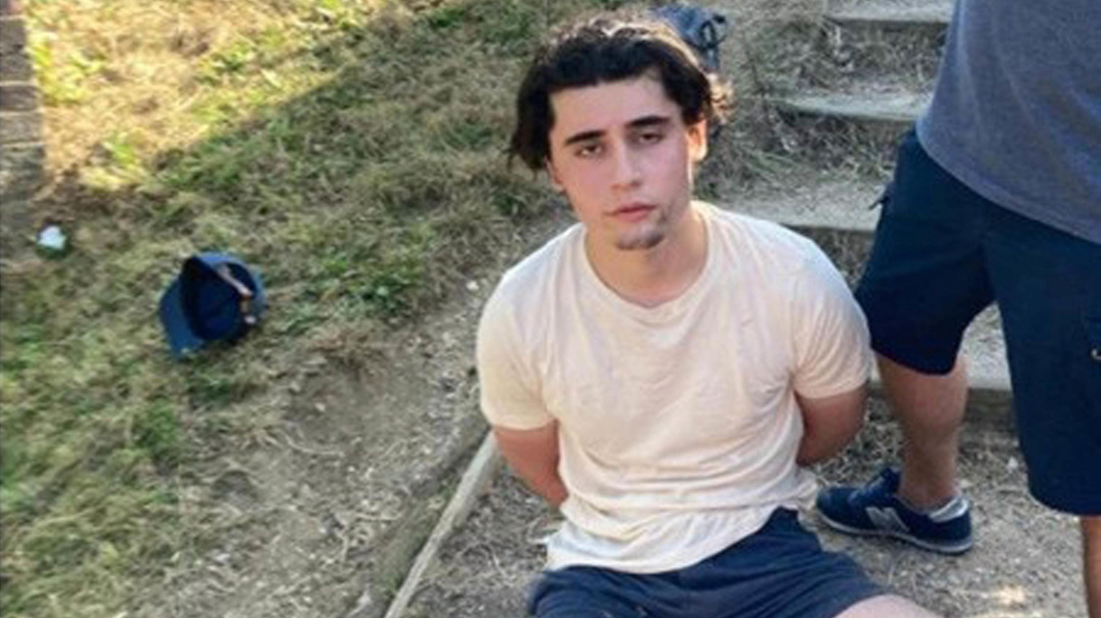 Daniel Khalife, wearing a white t-shirt, is pictured sitting on the ground outside with his hands behind his back after his arrest