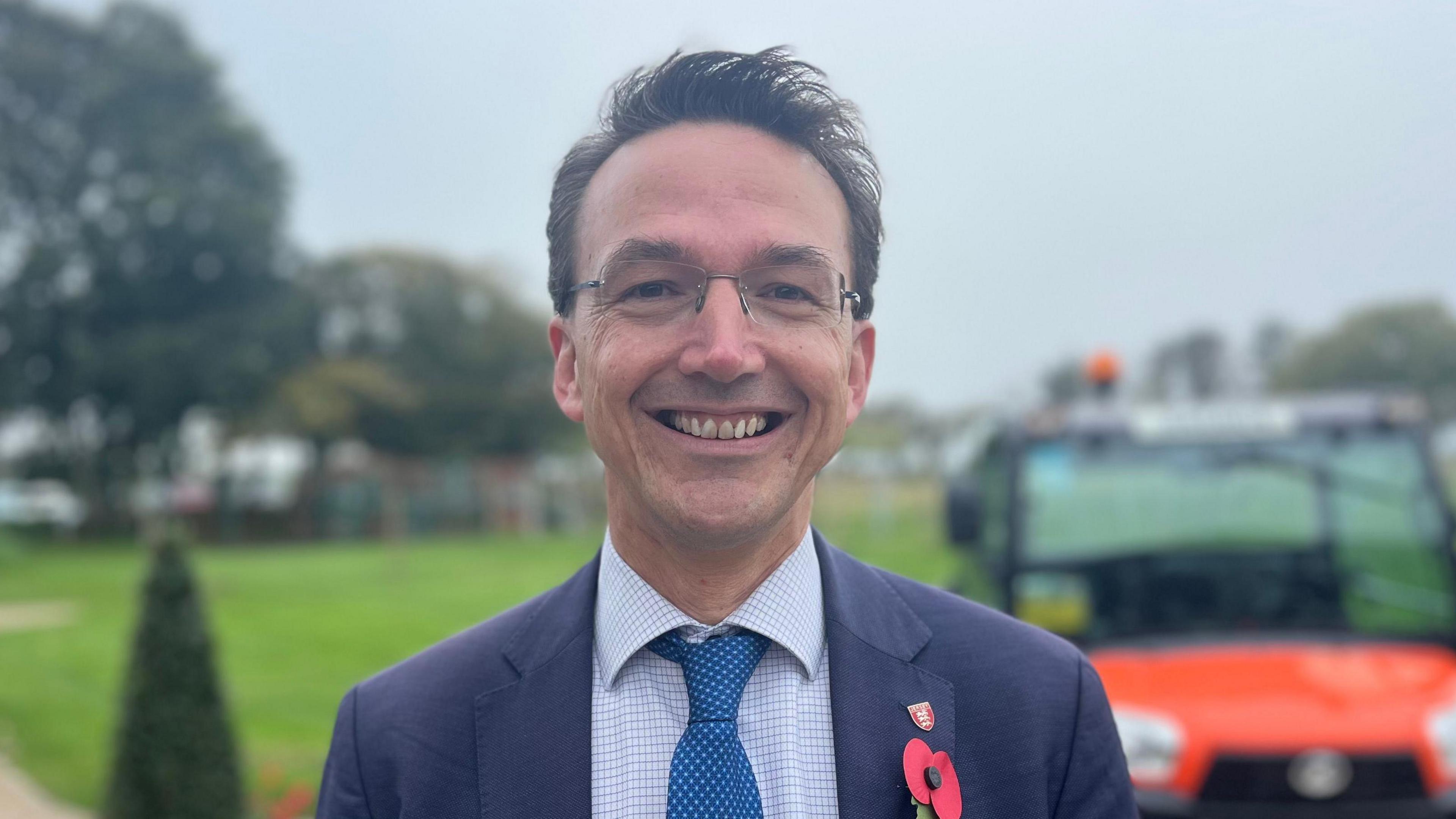 Sustainable Economic Development Minister Deputy Kirsten Morel smiling in park. He is wearing rimless glasses, a navy blue blazer with a poppy and a Jersey badge on it, a blue chequered shirt and dark blue tie. There is an orange vehicle behind him.