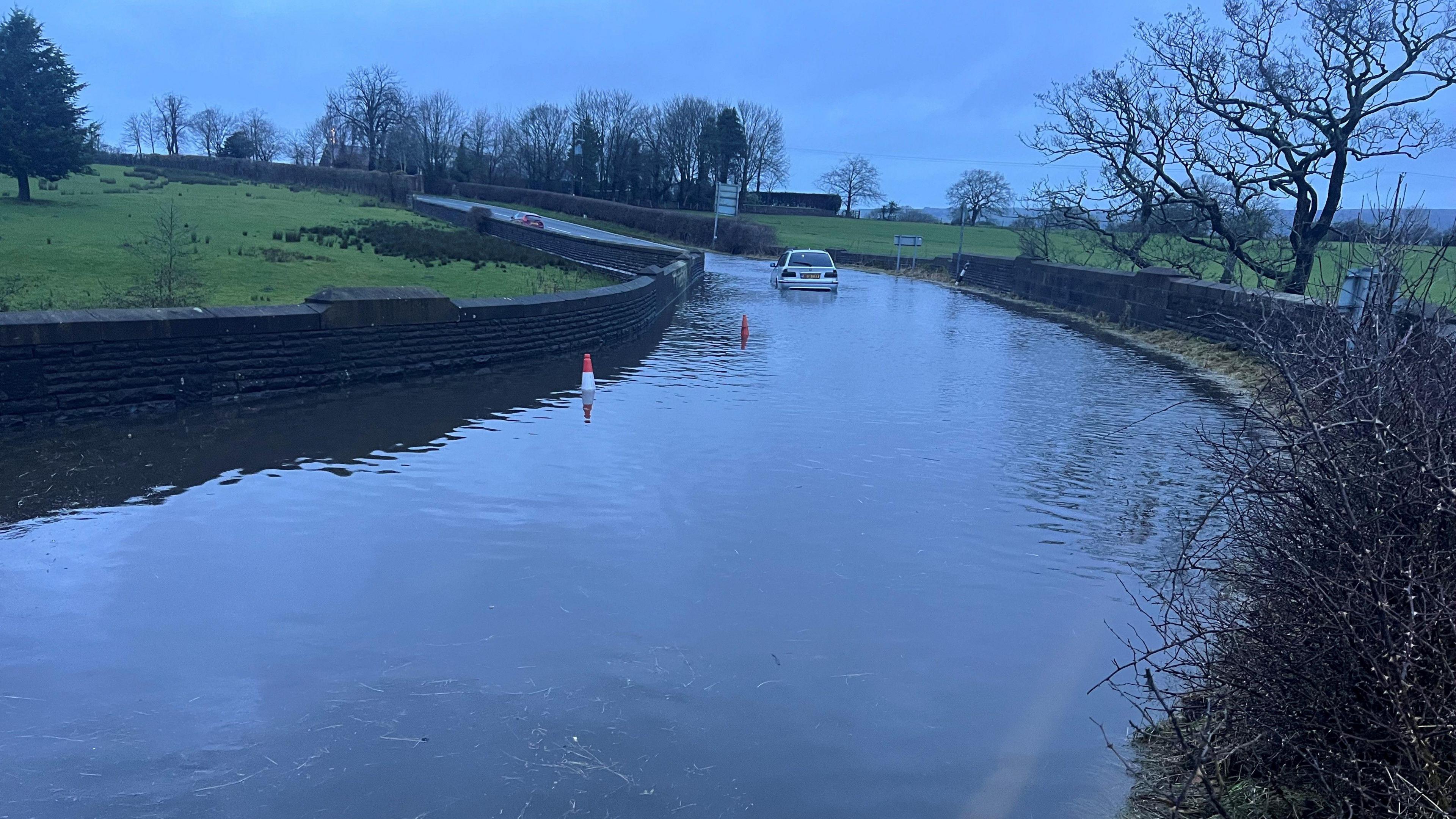 A stranded white car on a section of flooded road surrounded by green fields