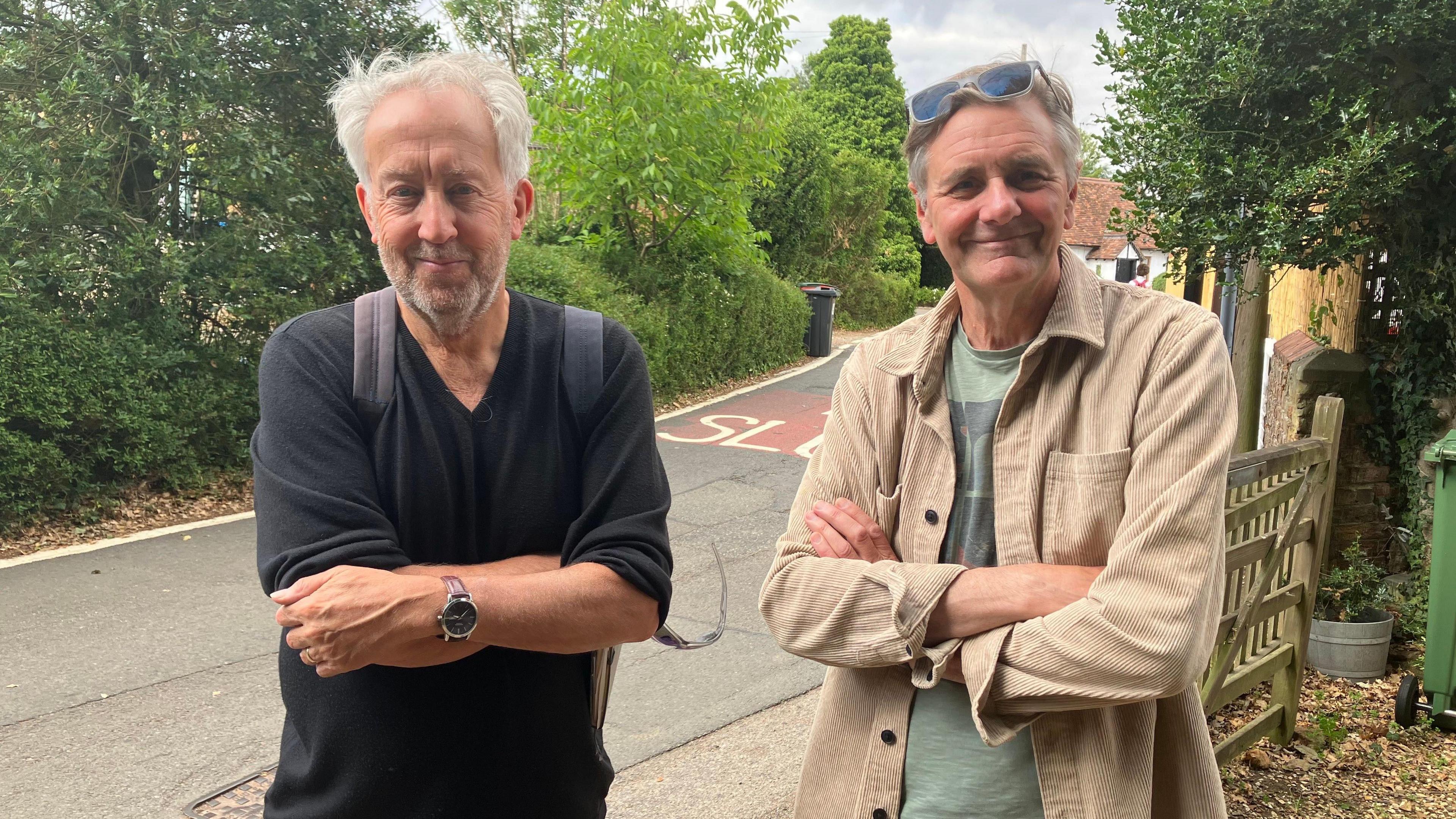 A picture of two men, stood next to a road. On the left is Conservative councillor David Hopkins, who has a black t-shirt on, has his arms folded and has a rucksack on his back. On his right is campaigner Trevor Hutton who also has his arms folded, but is wearing a light brown jacket over the top of a green t-shirt. He has sunglasses on his head.