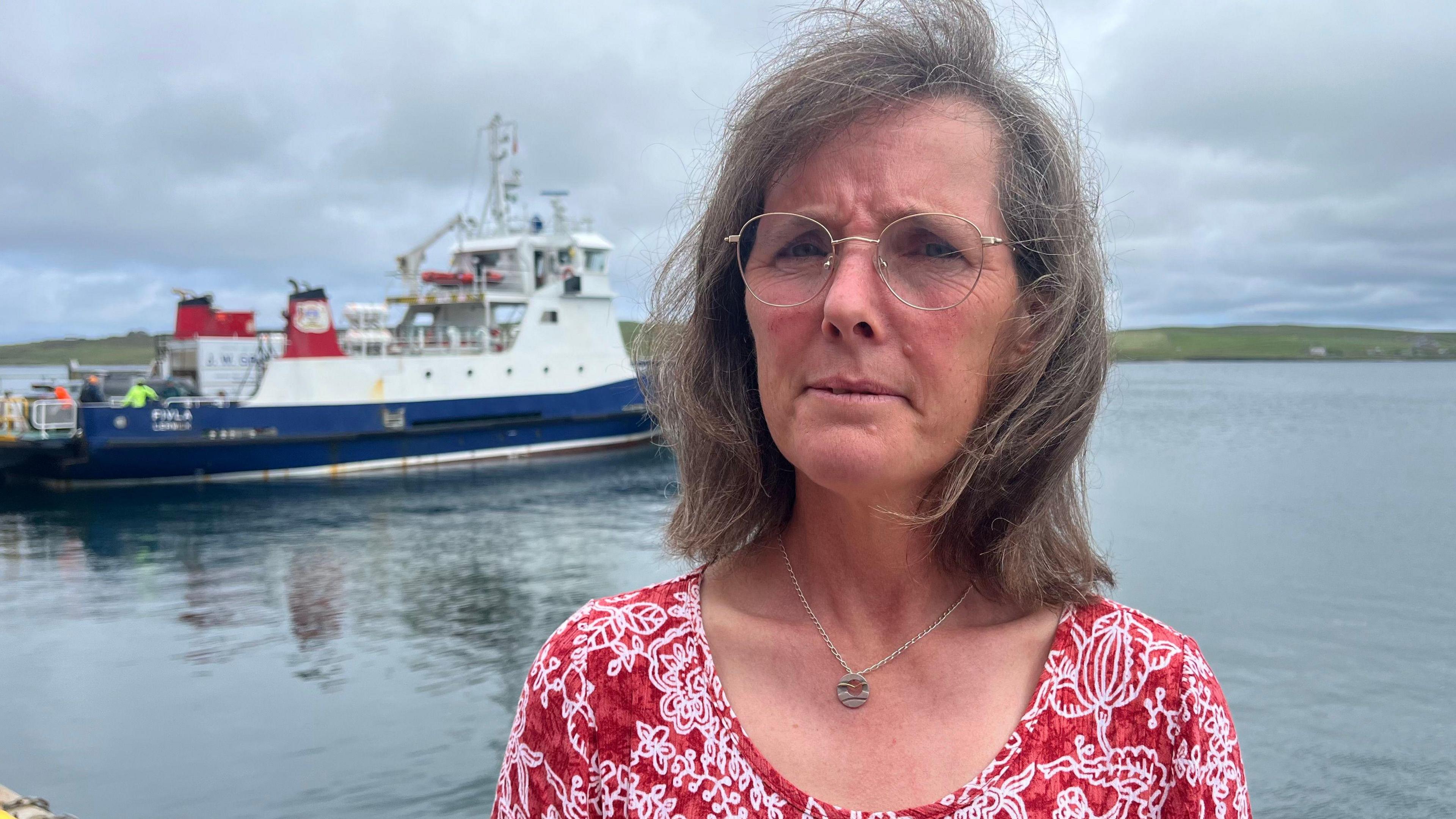 Close-up of a person standing by a harbour wearing round glasses and a red patterned top, with a ferry boat docked on calm water in the background under a cloudy sky.