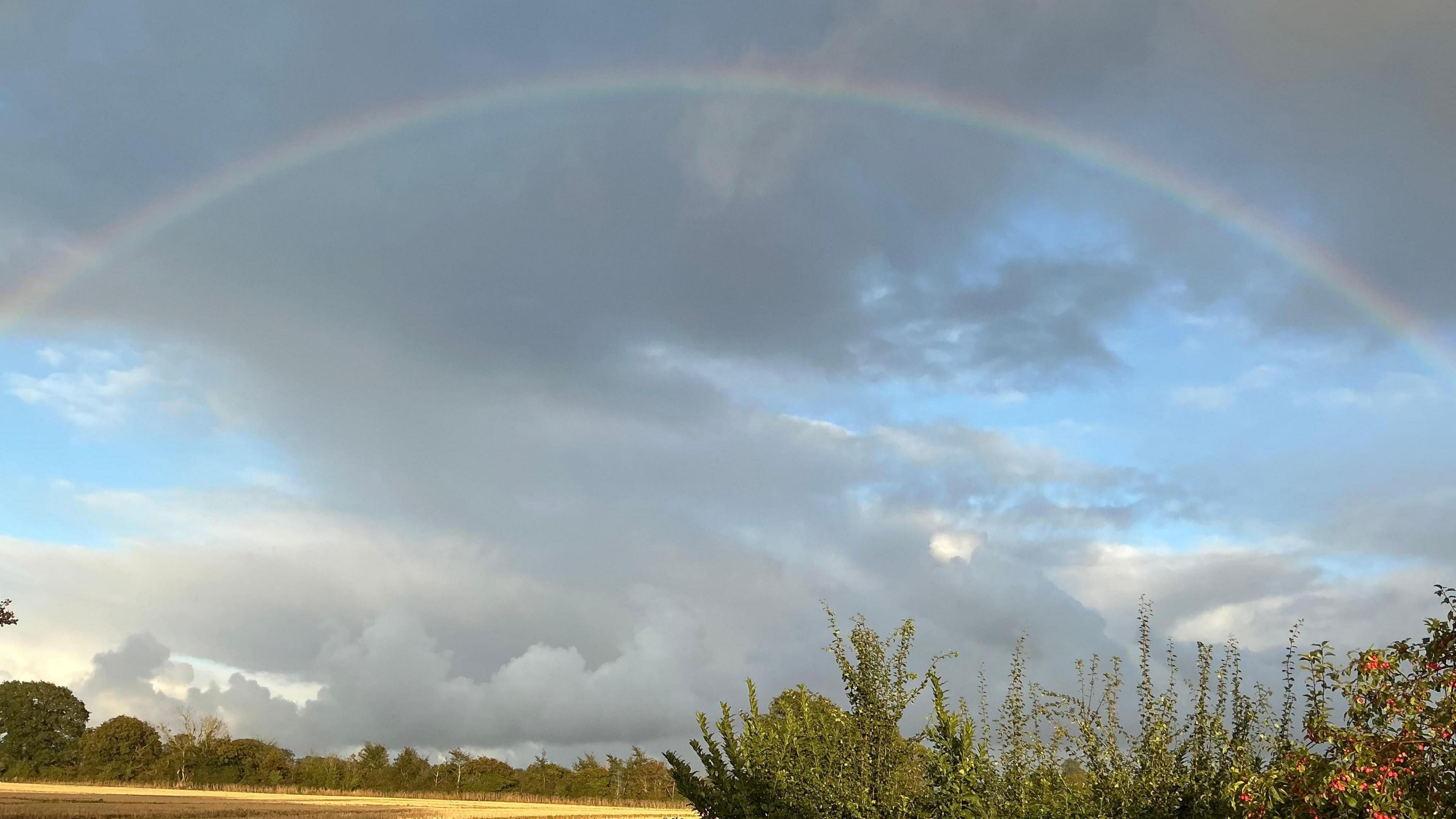 A large rainbow above a rural field. Thick grey clouds can be seen above the rainbow. 