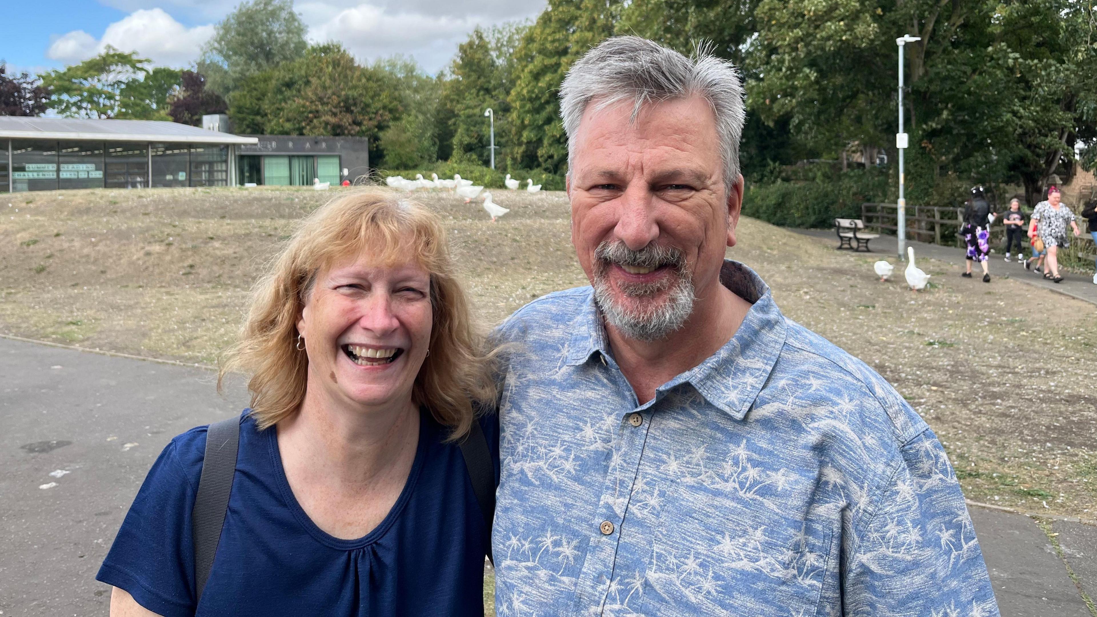 Nigel Lambert, 60 and his wife Julie, 60, stand in a park with geese visible behind them on a slight grassy mound. Julie has shoulder length blonde hair and is smiling with a V-necked blue top and a leather shoulder strap over her right shoulder. Nigel Lambert has short grey hair which is a bit spikey he has a neat grey beard and moustache and is wearing a blue and white patterned shirt and has his right arm around his wife's shoulder.