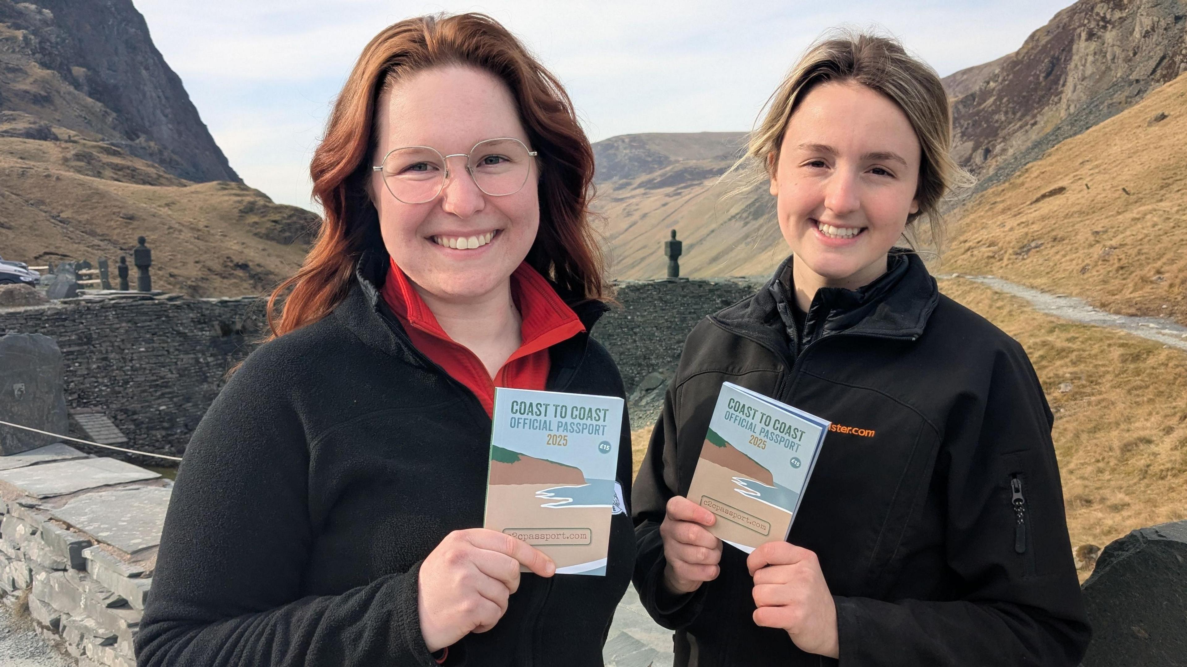 Two women hold up Coast to Coast official passports at a slate mine. The landscape has steep hills.