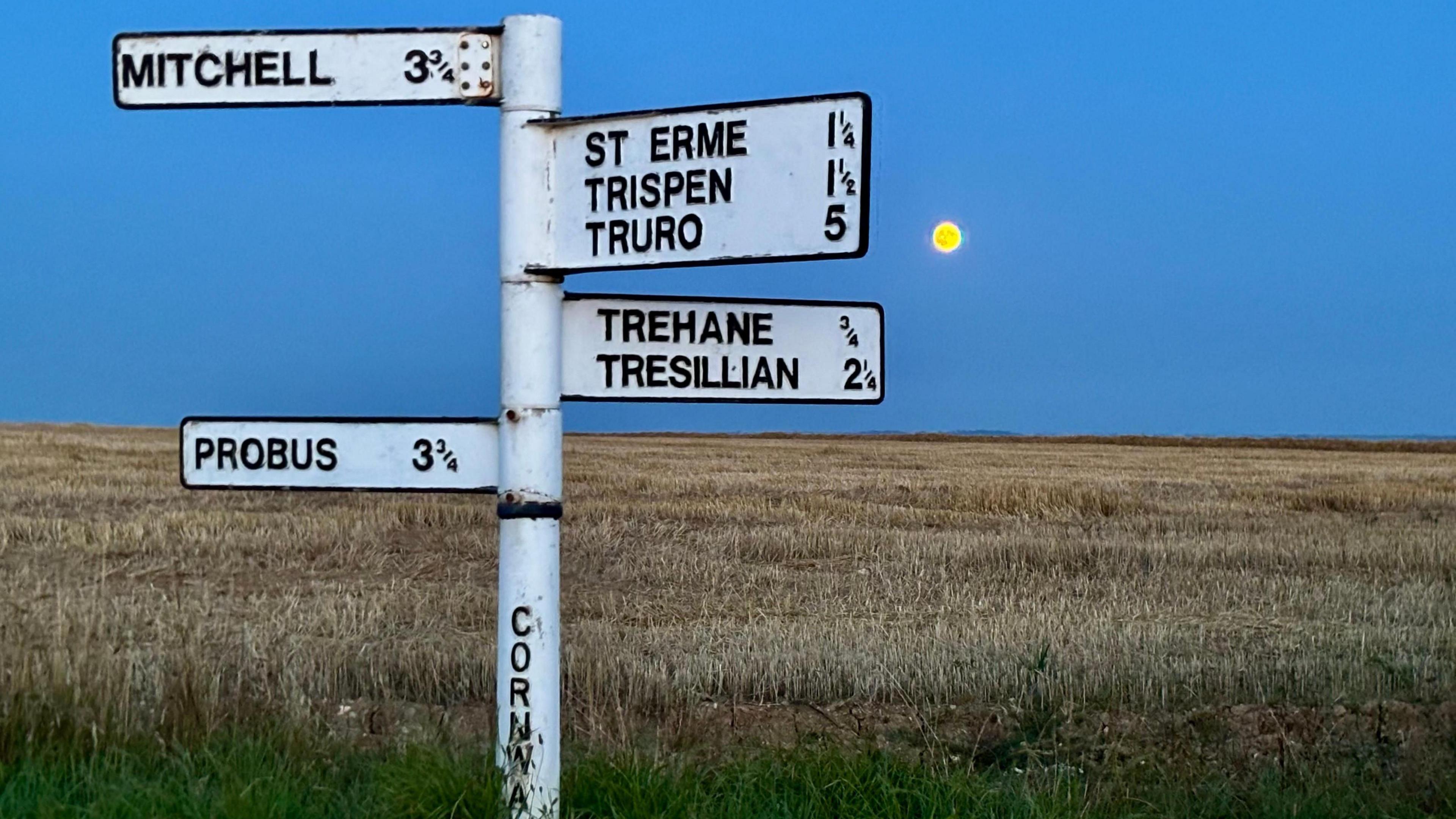 White rural signpost in St Erme, Cornwall, standing beside a harvested field at dusk, with a small golden full moon low on the horizon in the background.
